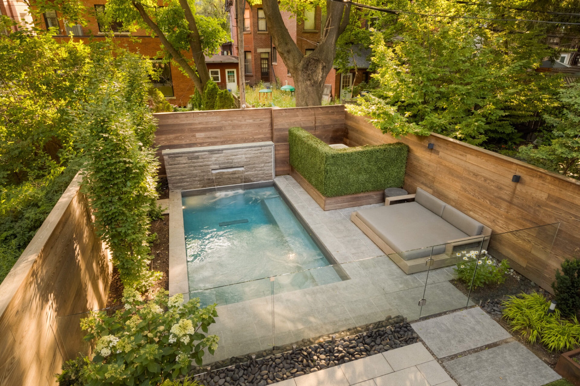 A serene backyard features a small pool, waterfall, and lounge area, surrounded by lush greenery and wooden fencing, creating a private oasis.