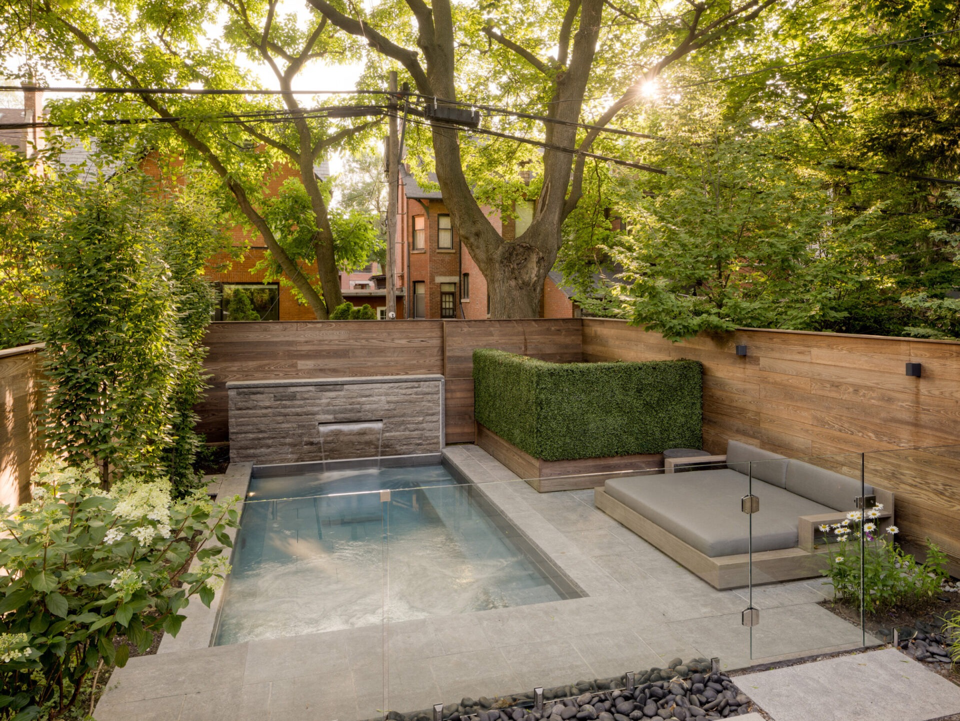 A serene backyard features a small pool, modern lounge chair, lush greenery, and wooden fencing, offering a private retreat under dappled sunlight.