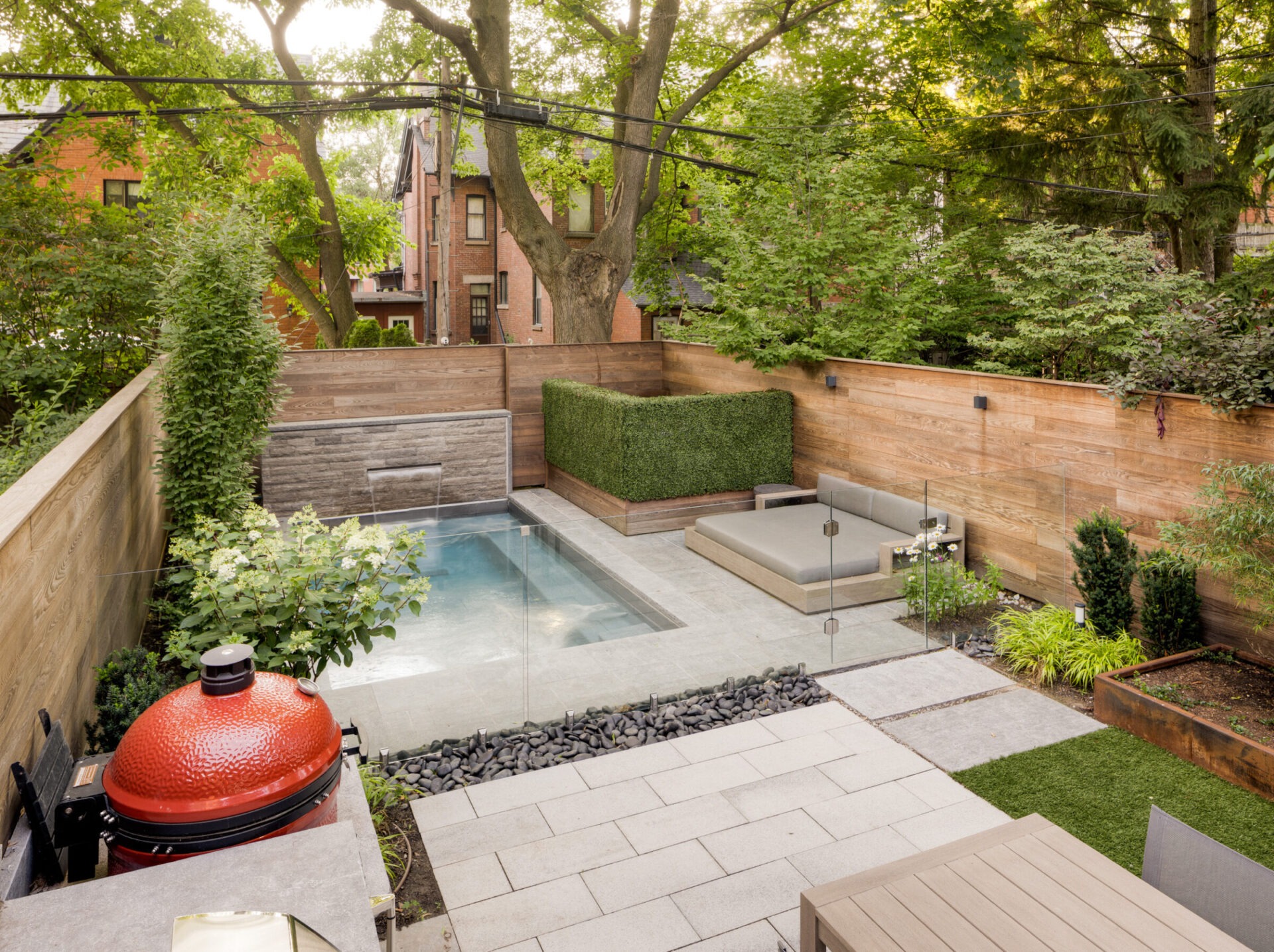 Modern backyard with a small pool, patio, and red grill. Surrounded by greenery and wood fencing. Brick houses visible in background.