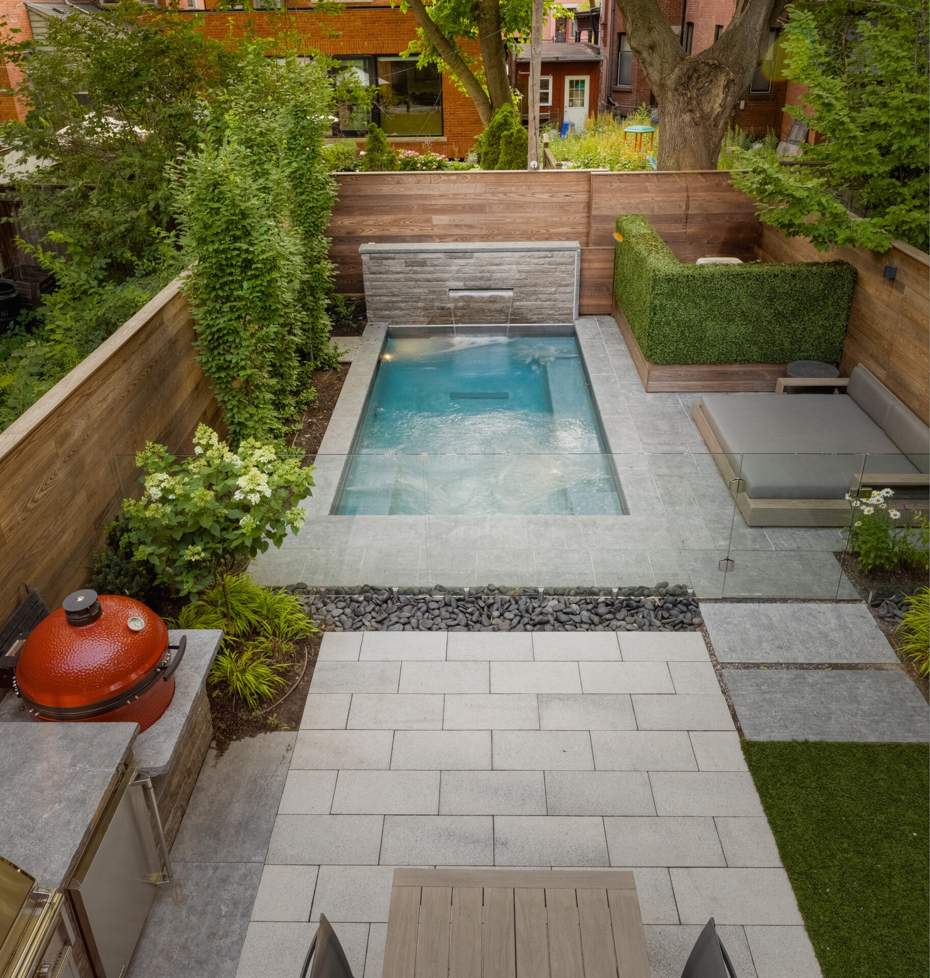 A modern backyard features a small pool, glass fencing, lounge seating, and a red outdoor grill surrounded by greenery and brick buildings.