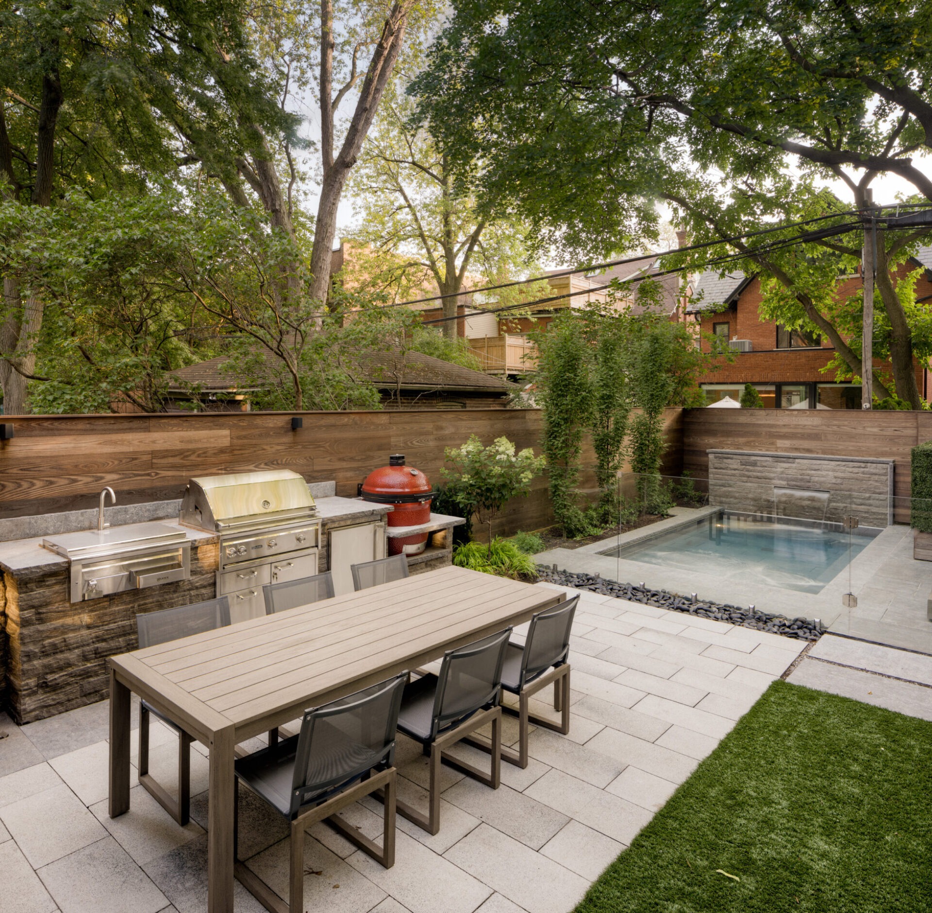 Outdoor patio features a modern barbecue grill, dining table, chairs, and a small pool surrounded by trees and wooden fencing.