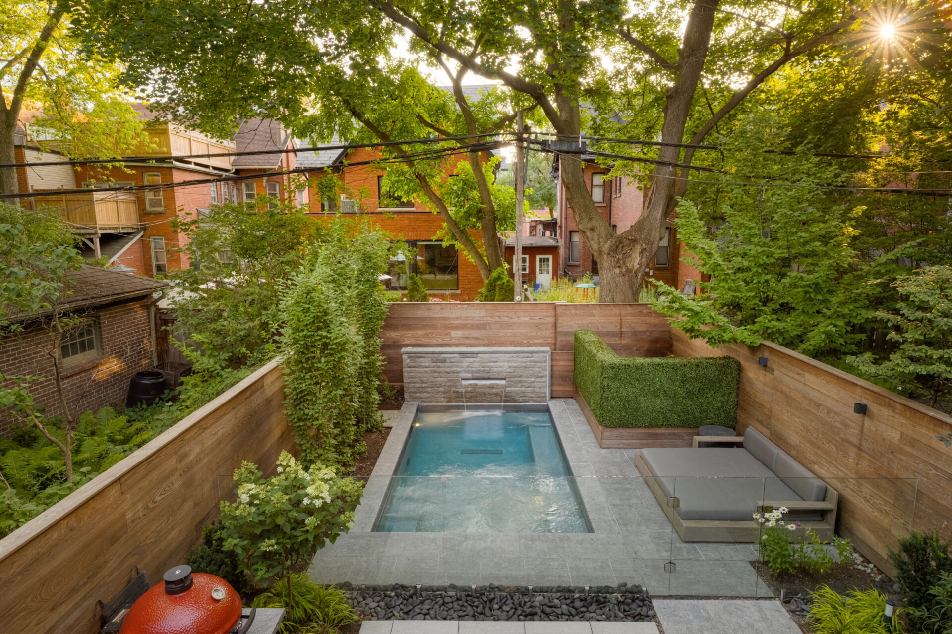 A backyard features a small pool, lounge area, and greenery, surrounded by wooden fences and brick houses in a residential neighborhood.