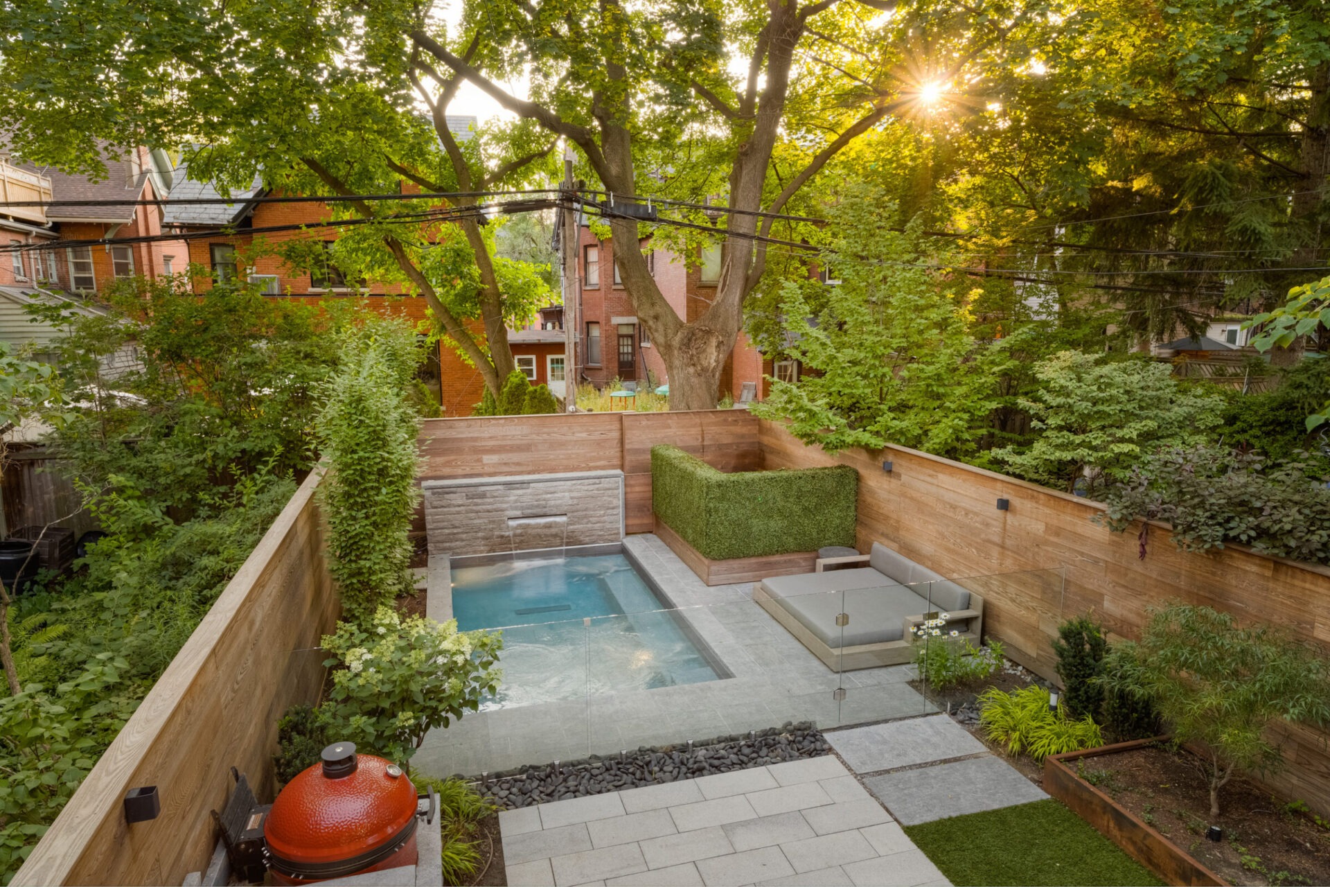 A serene backyard features a small pool, lush greenery, and comfortable seating. Red brick buildings and leafy trees are in the background.