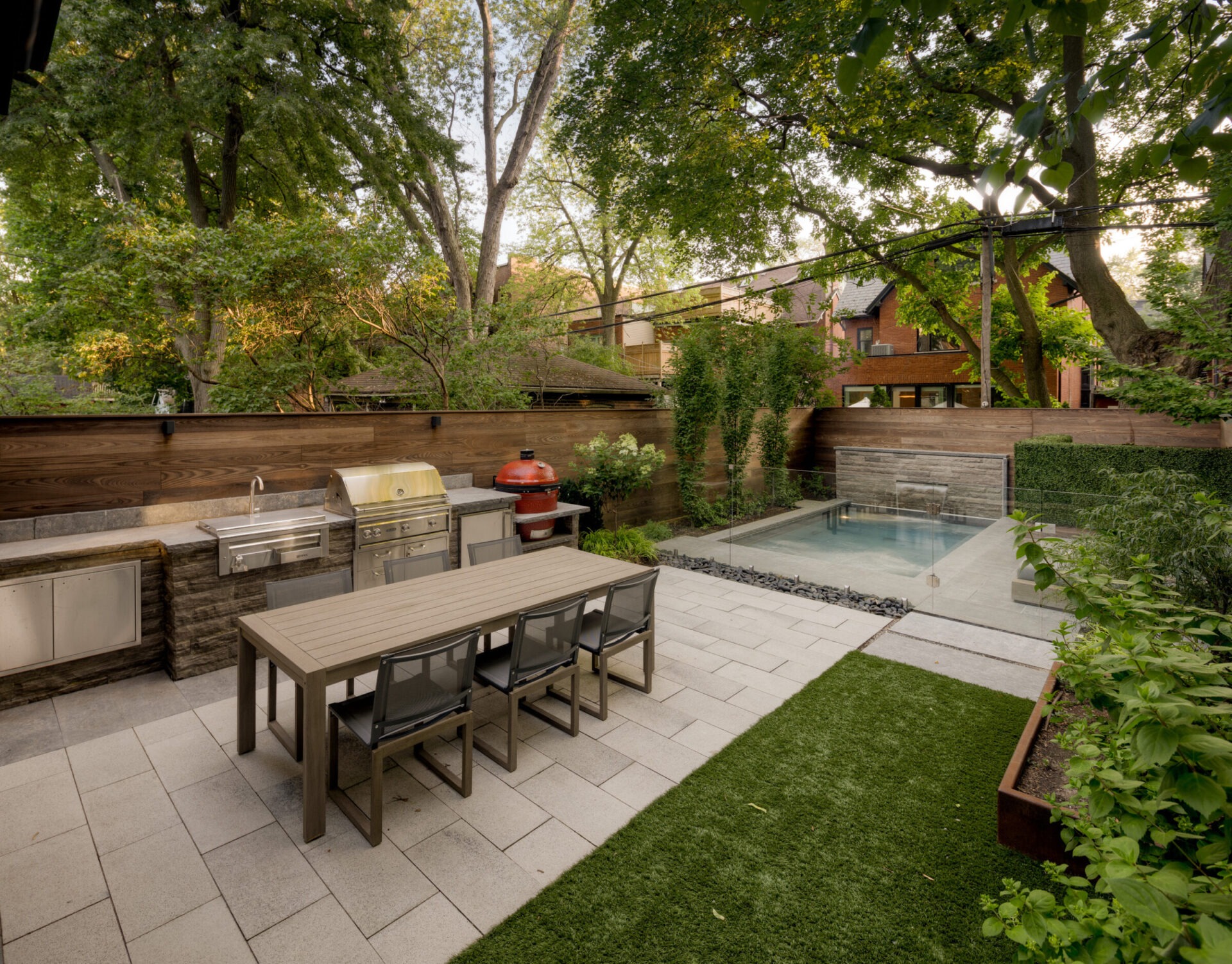 A modern backyard features an outdoor kitchen with grill, dining table, and a small pool, surrounded by trees and greenery.