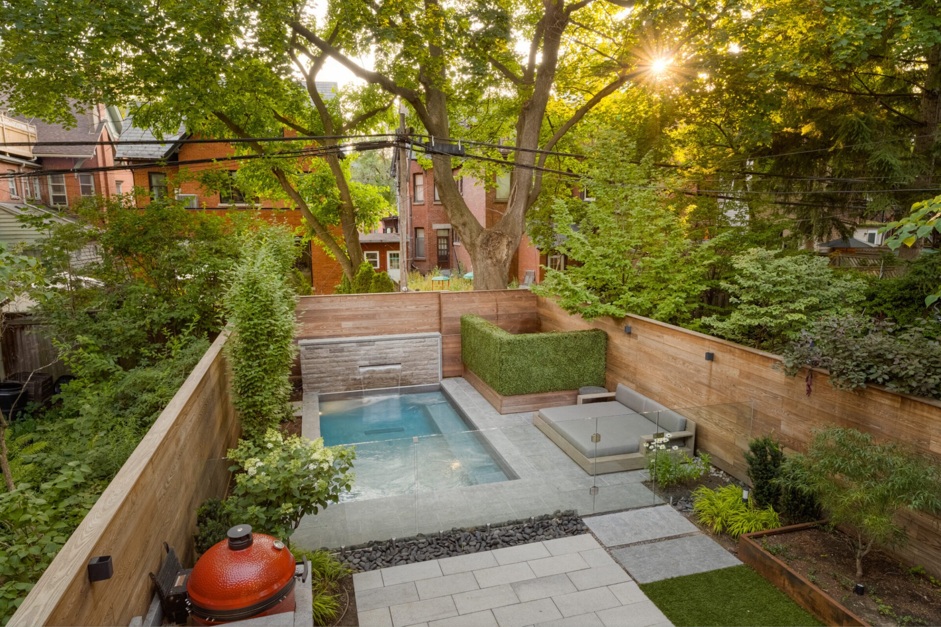 A serene backyard features a small pool, seating area, lush greenery, and a grill, with sunlight filtering through the surrounding trees.