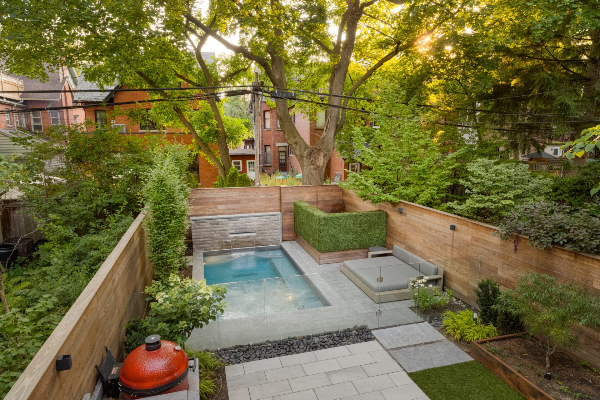 A tranquil backyard features a small pool, seating area, grill, and lush greenery, with brick houses visible in the background.