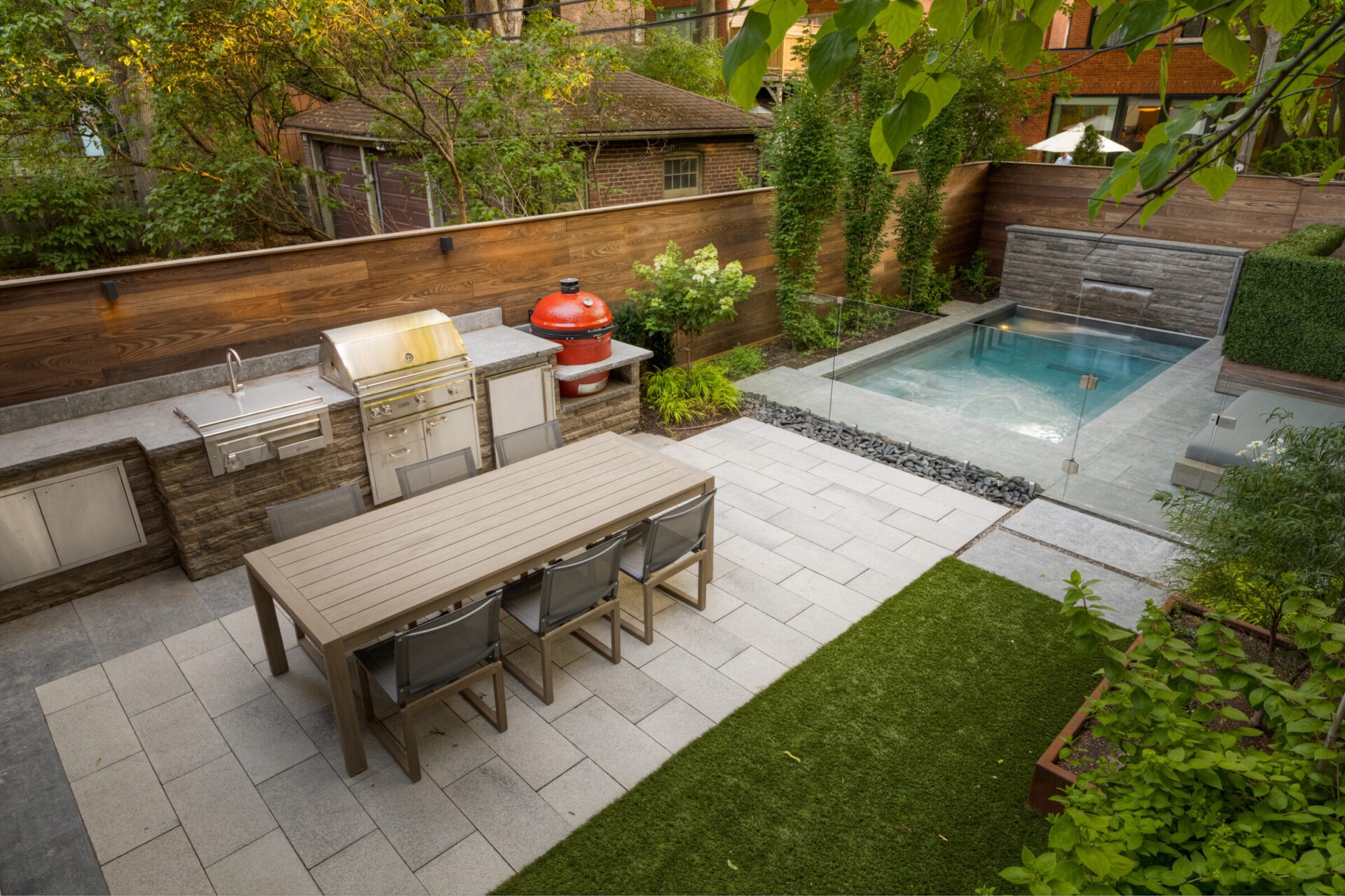 Modern backyard with a dining table, outdoor kitchen, and small pool, surrounded by lush greenery and wooden fencing. Peaceful urban retreat.