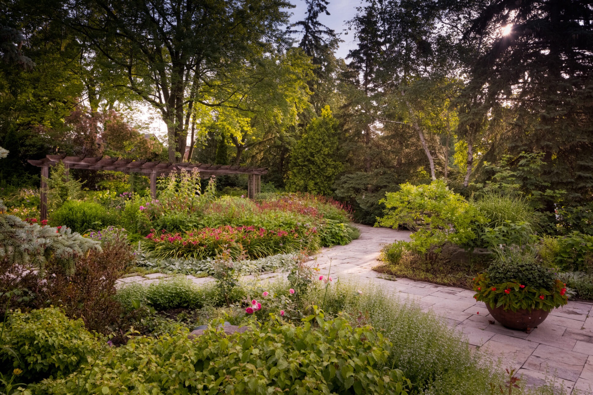 Lush garden with colorful flowers, stone pathway, and wooden pergola set amidst tall, green trees under a clear sky, creating a tranquil atmosphere.