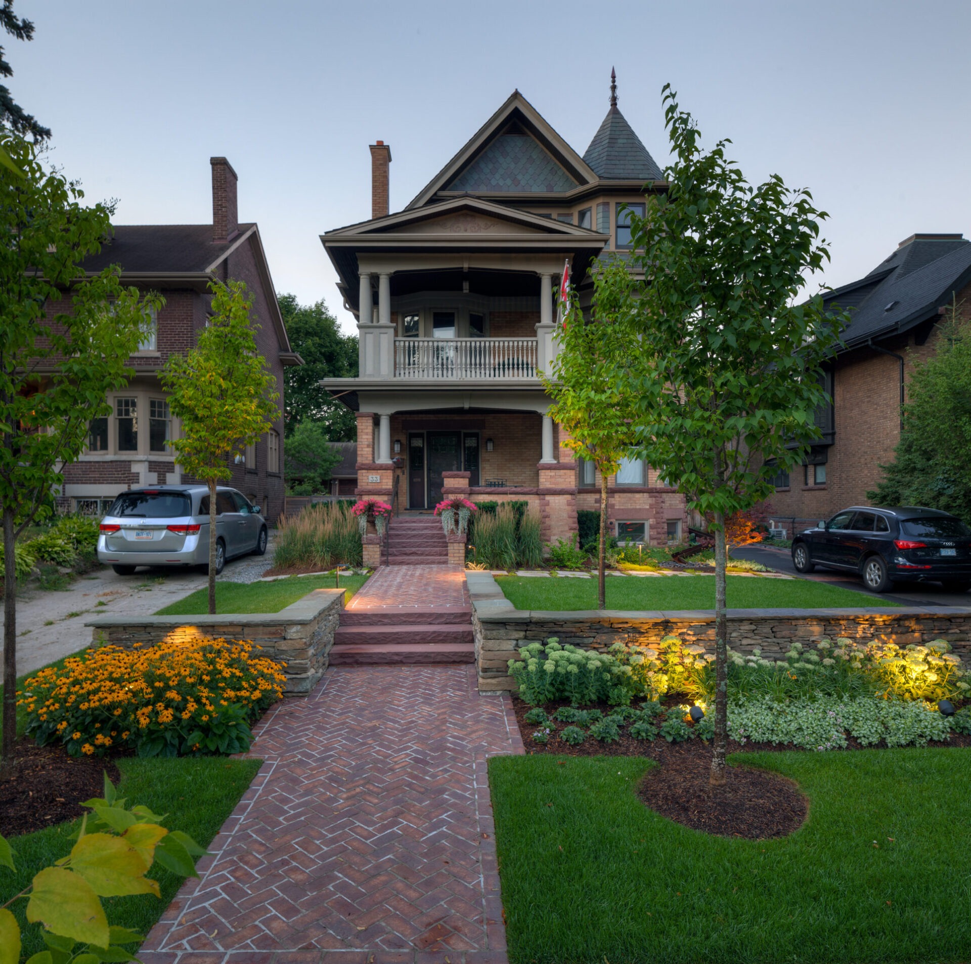 A brick path leads to a two-story Victorian house with well-maintained gardens, surrounded by cars and neighboring homes, under a clear sky.