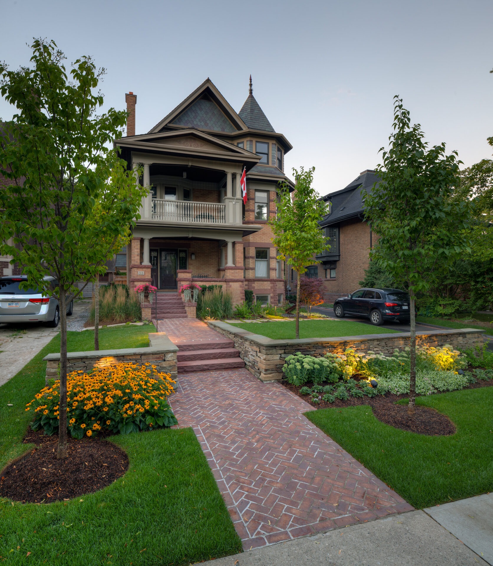 Victorian-style house with brick path, garden, and two cars parked in the driveway. Canadian flag displayed on the upper balcony.