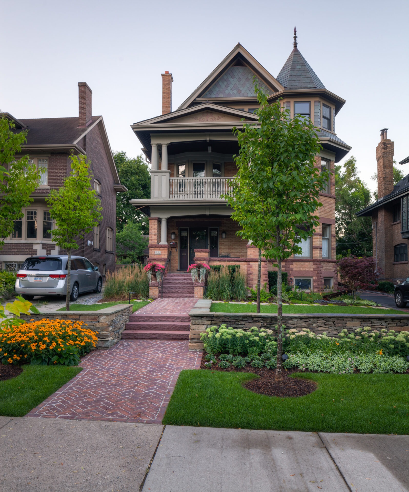 Victorian-style house with turret and covered porch, surrounded by lush gardens and brick walkway. Adjacent buildings partly visible in suburban setting.