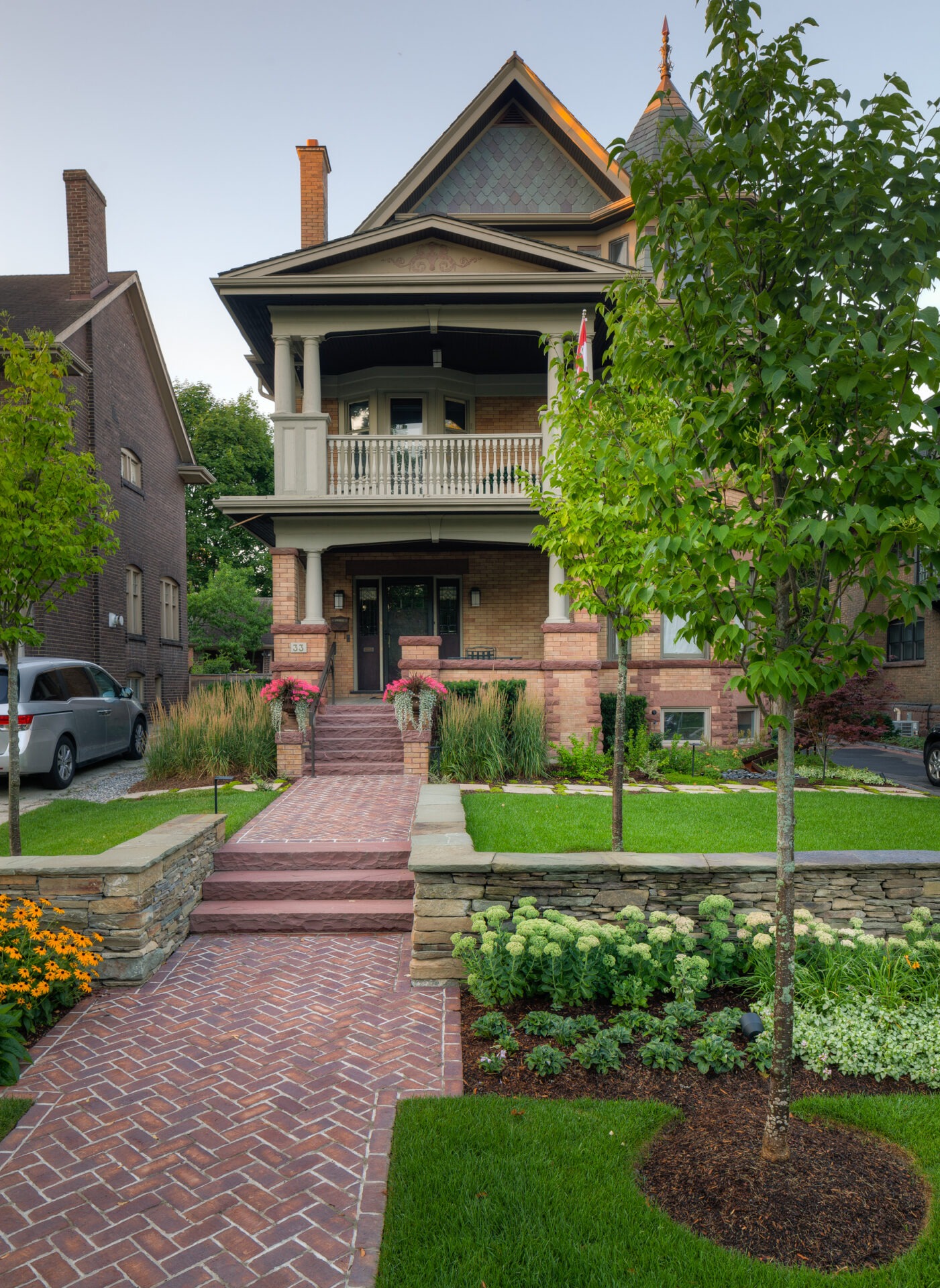 A well-maintained brick Victorian house with a porch, surrounded by lush greenery and flowers, alongside a minivan parked in the driveway.