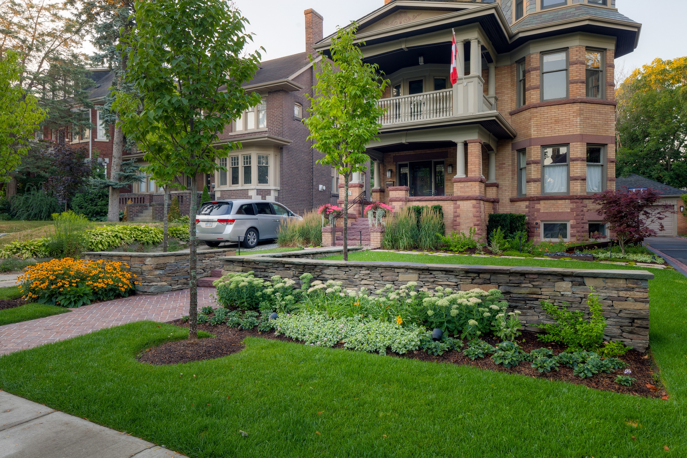 A large Victorian-style house with a tower, Canadian flag, lush garden, brick walkway, nearby parked cars, lined by green lawns and trees.