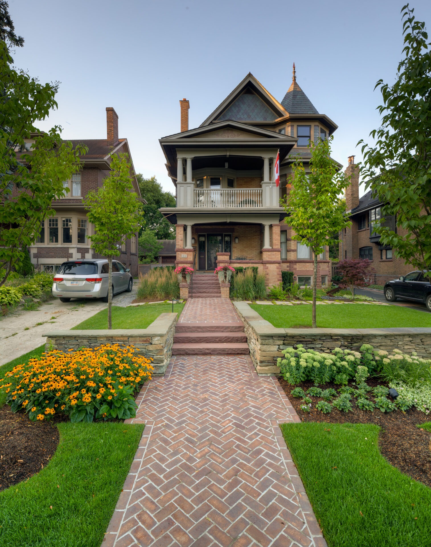 A large Victorian-style house with a tower, Canadian flag, lush garden, brick walkway, nearby parked cars, lined by green lawns and trees.