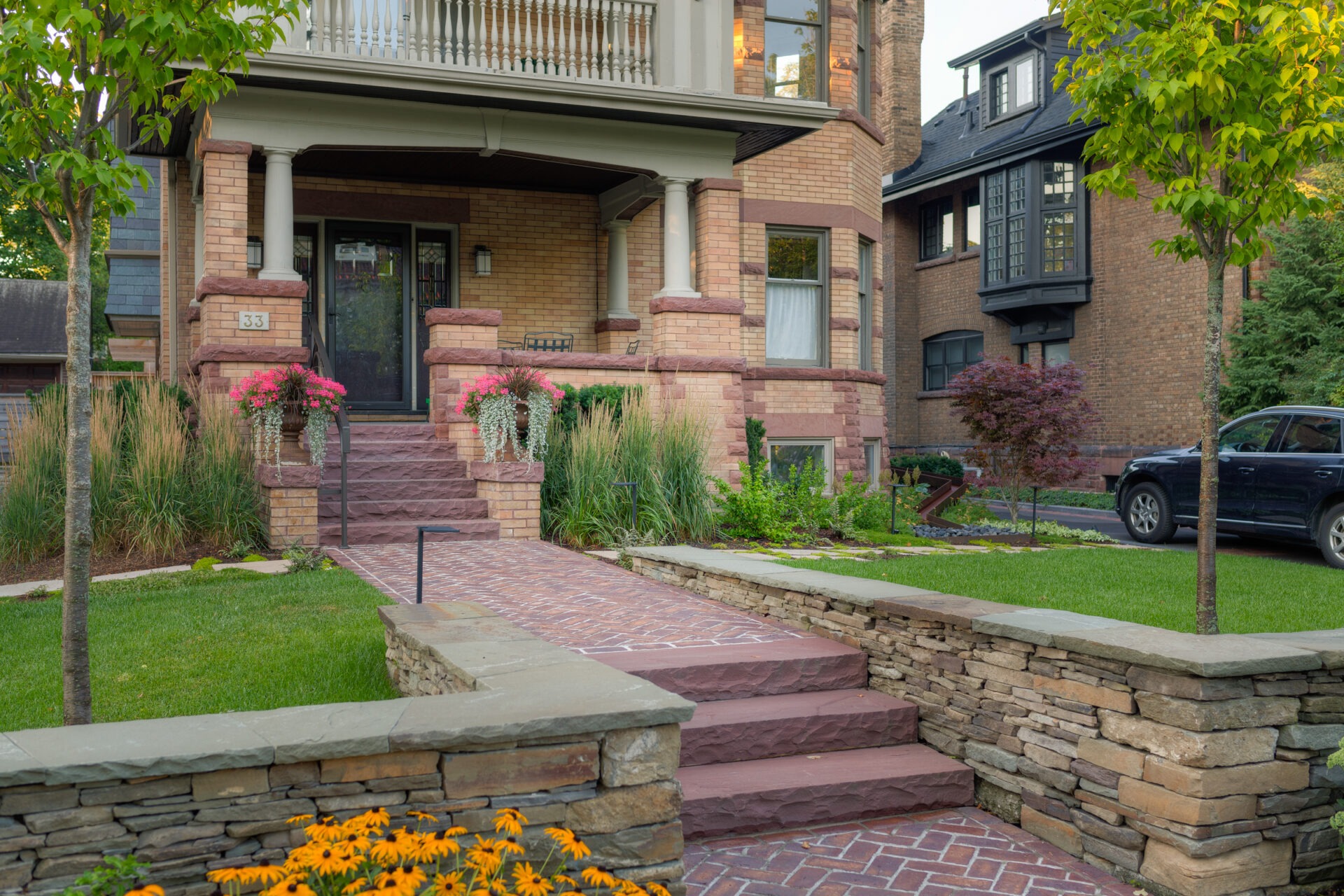 Charming brick house with a well-manicured garden and pathway. A vehicle is parked nearby. Vibrant flowers and greenery enhance the scene.