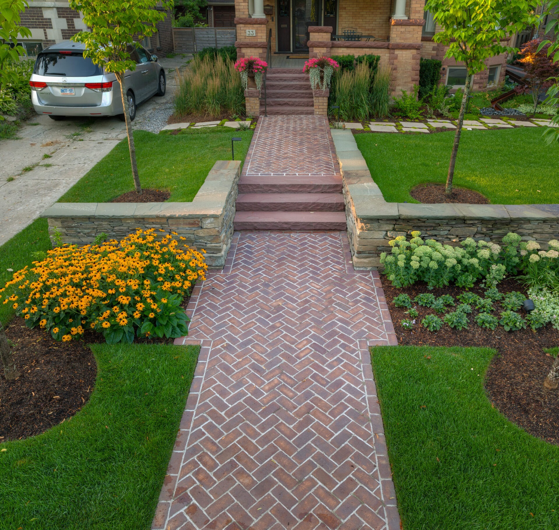 A well-maintained front yard features a herringbone brick path, vibrant flowers, lush grass, and a parked car near a brick house.