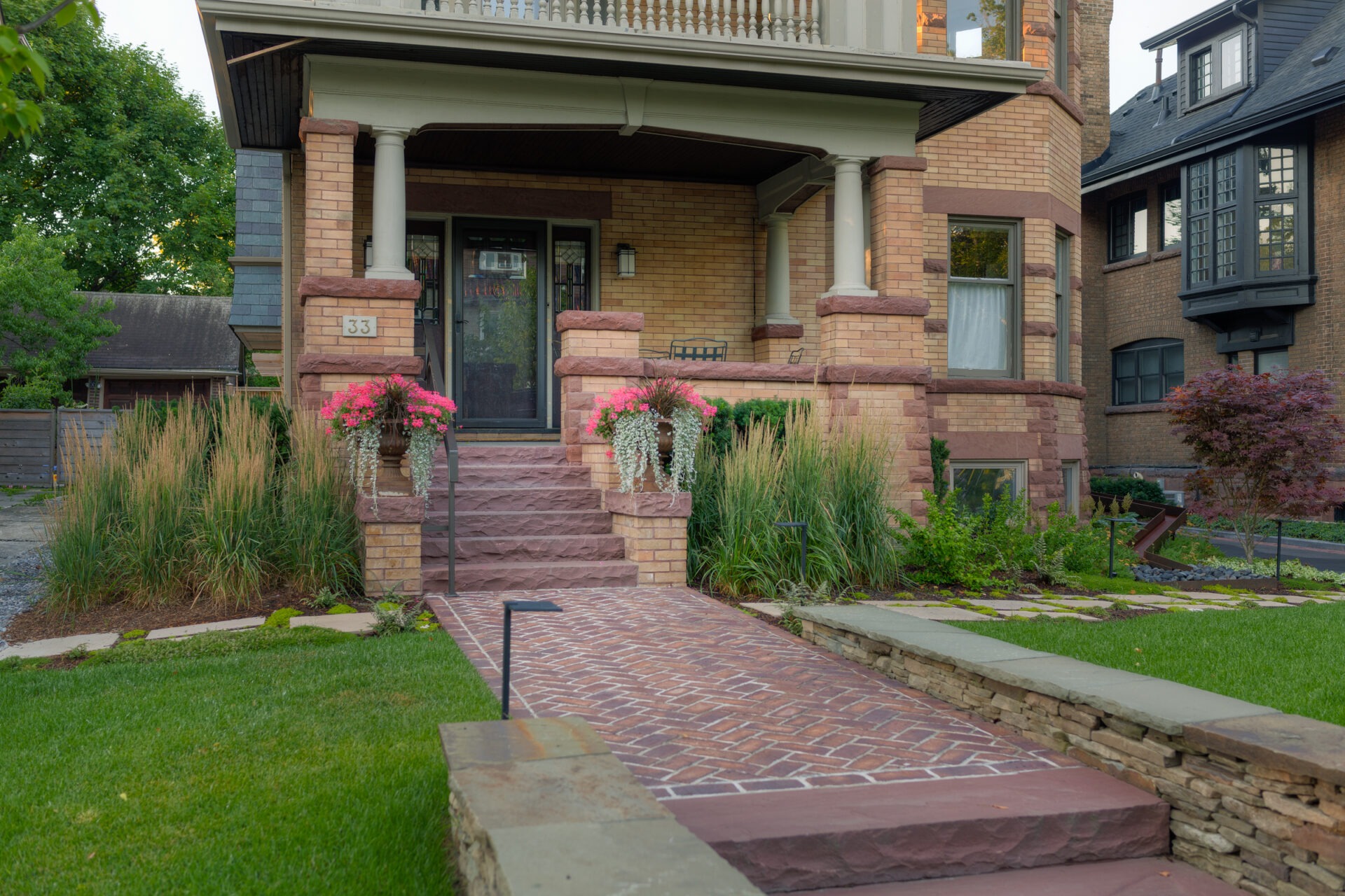 A charming brick house with a welcoming front porch, blooming flowers, manicured lawn, and a brick walkway leading to the entrance.