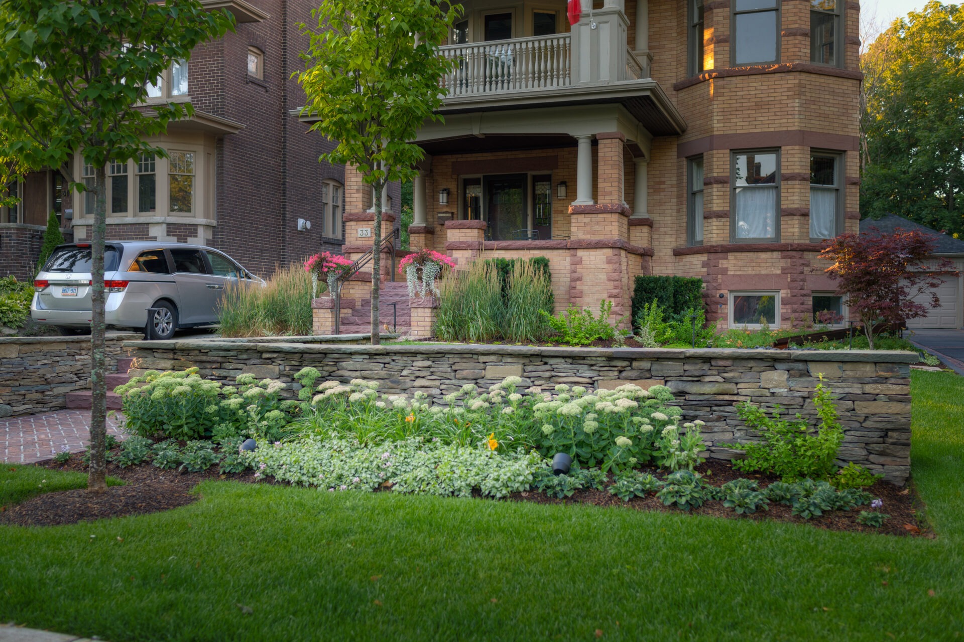 A well-maintained brick house with a green garden and stone wall, featuring a parked silver van in the driveway and lush landscaping.