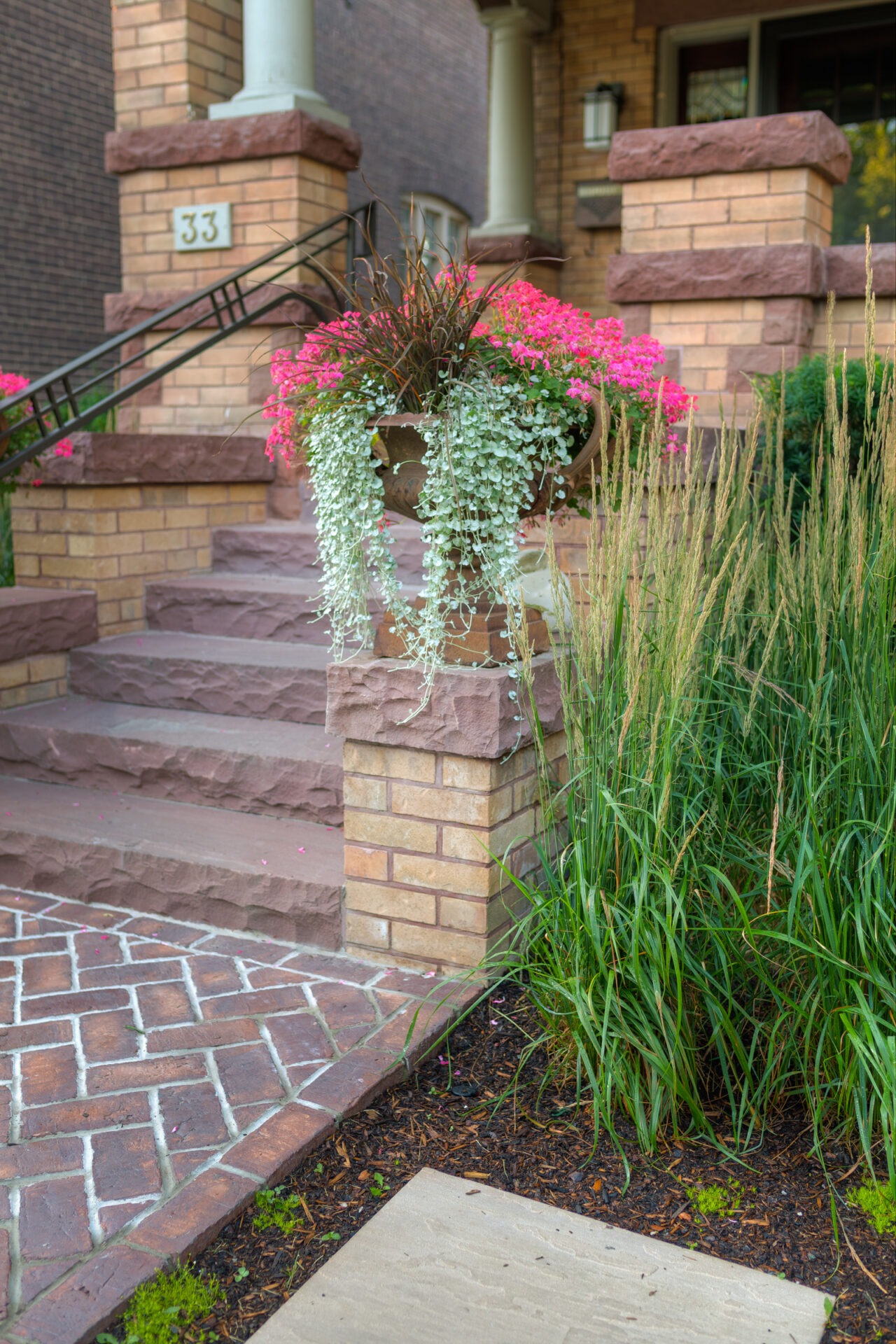 Brick steps with wrought iron railing, decorative pillars, and vibrant floral arrangement; lush green plants surround a charming entrance. House marked number 33.