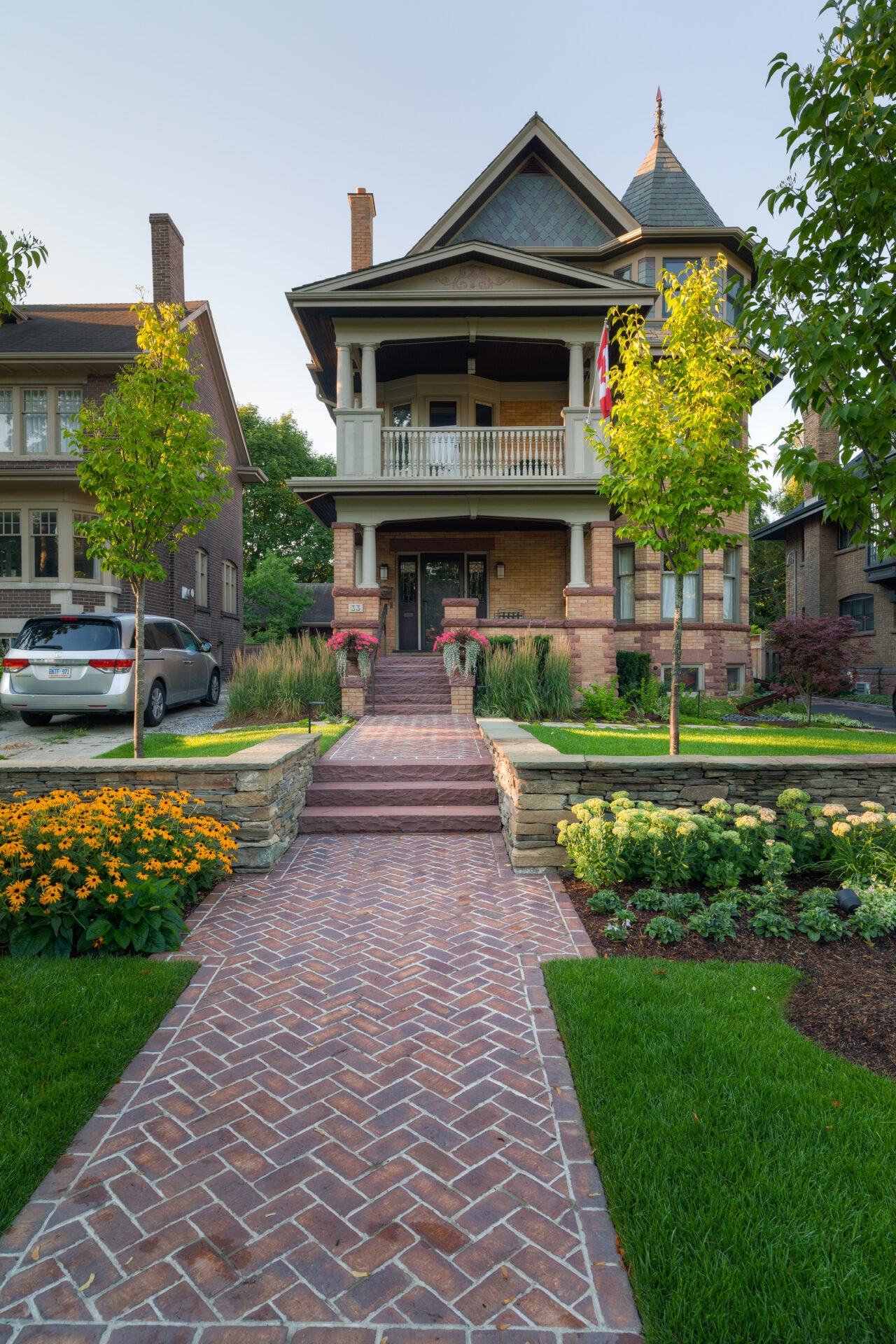 Victorian-style house with a gable roof and porch, surrounded by colorful gardens and stone pathways. Trees line the manicured lawn.