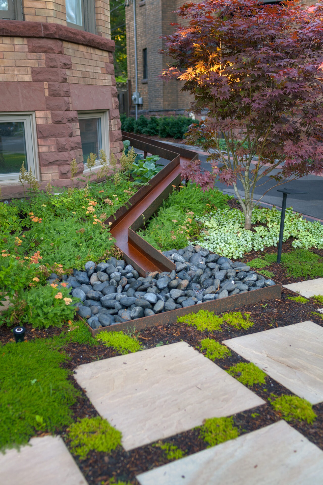 Brick building corner with a modern garden design, featuring a stone path, greenery, rusted metal water feature, and ornamental tree.