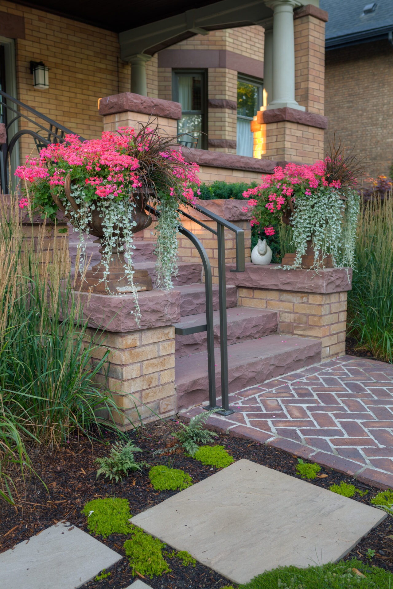 Stone steps lead to a brick house entrance, adorned with pink flowers and greenery, surrounded by a patterned walkway and lush plants.