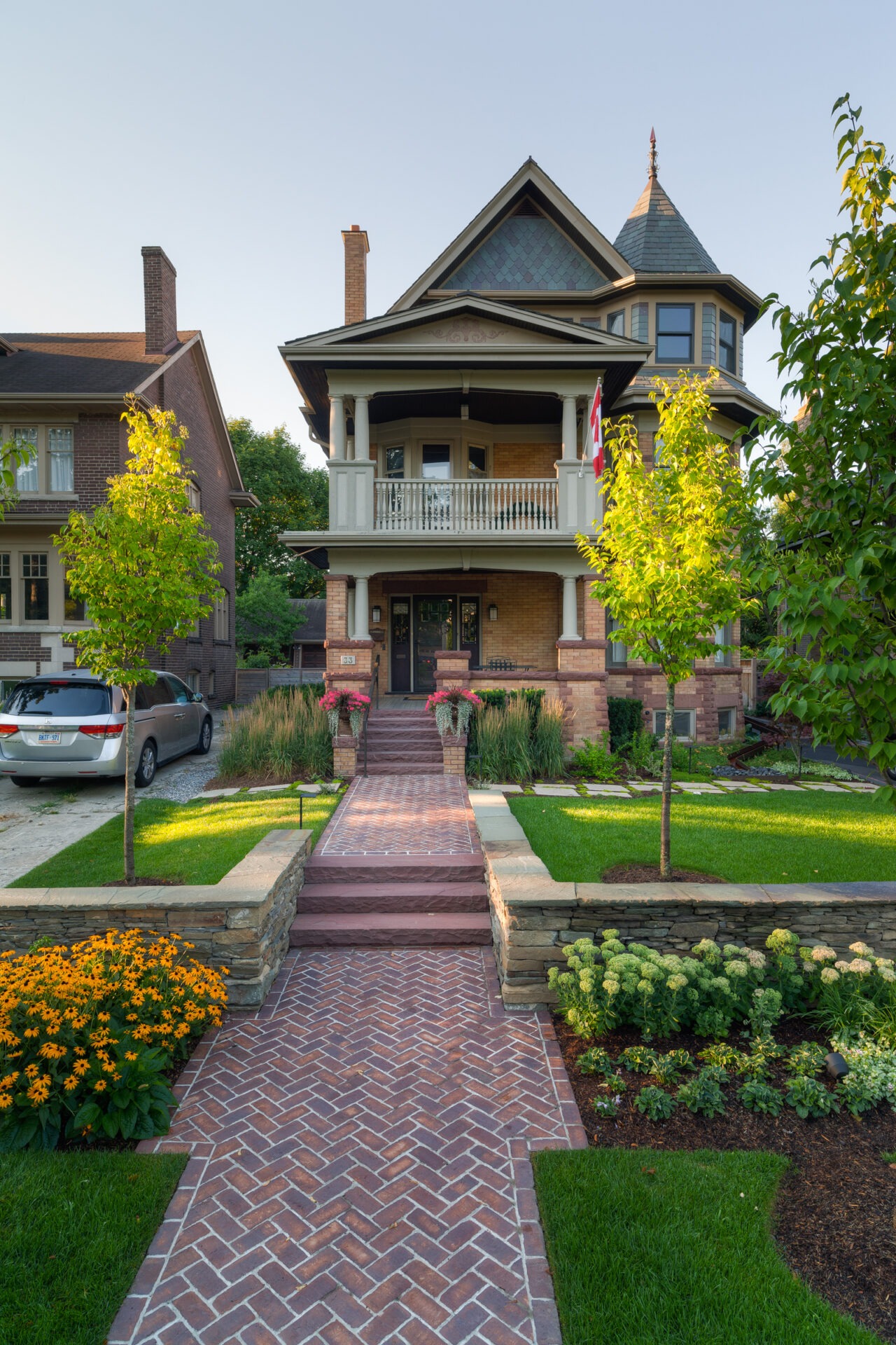 A charming, two-story brick house with a porch. Well-manicured garden, flowers, paved walkway, trees, and a driveway with a parked car.