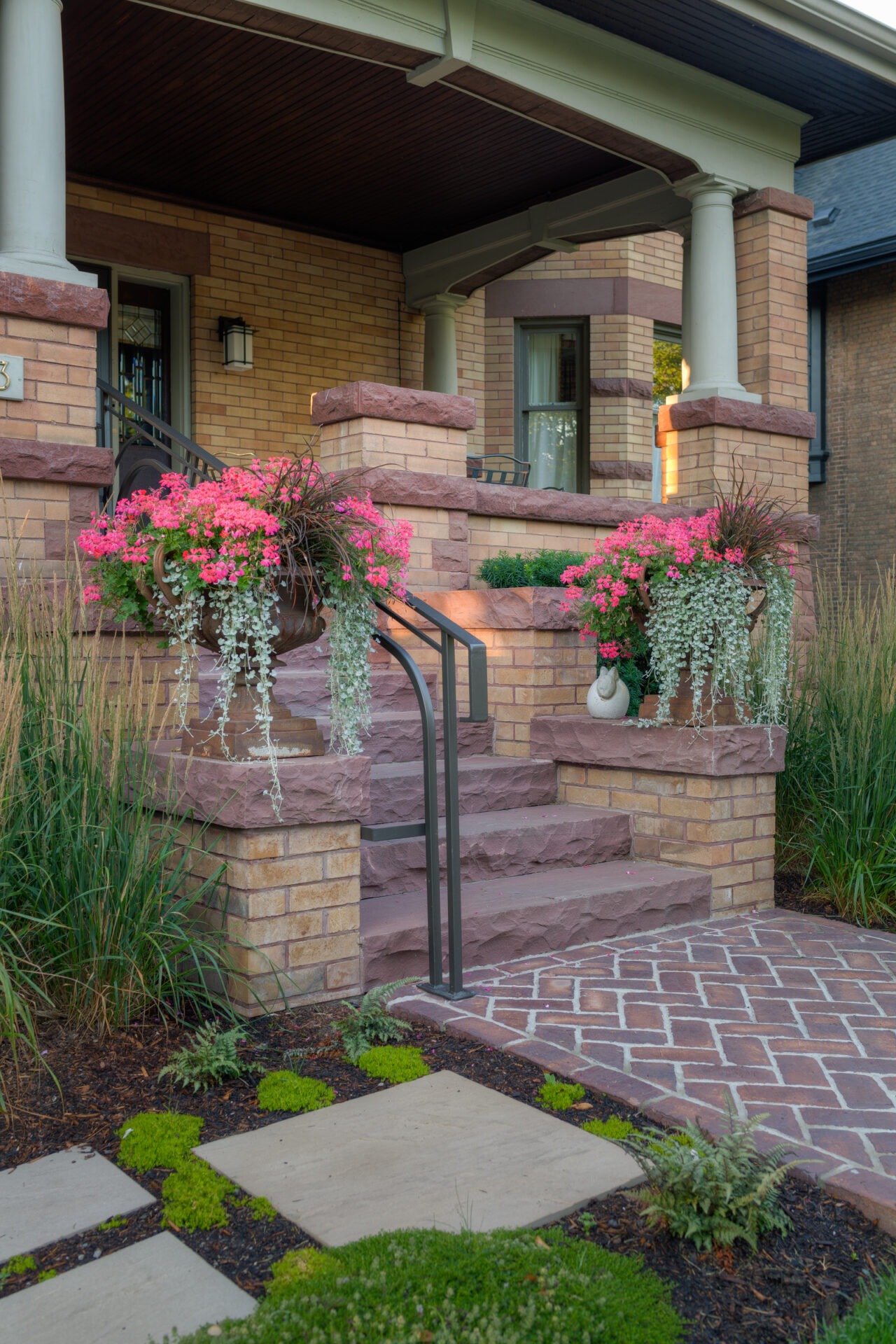 Brick porch with colorful flowers and greenery. Features stone steps and pillars, leading to a cozy, welcoming entrance.