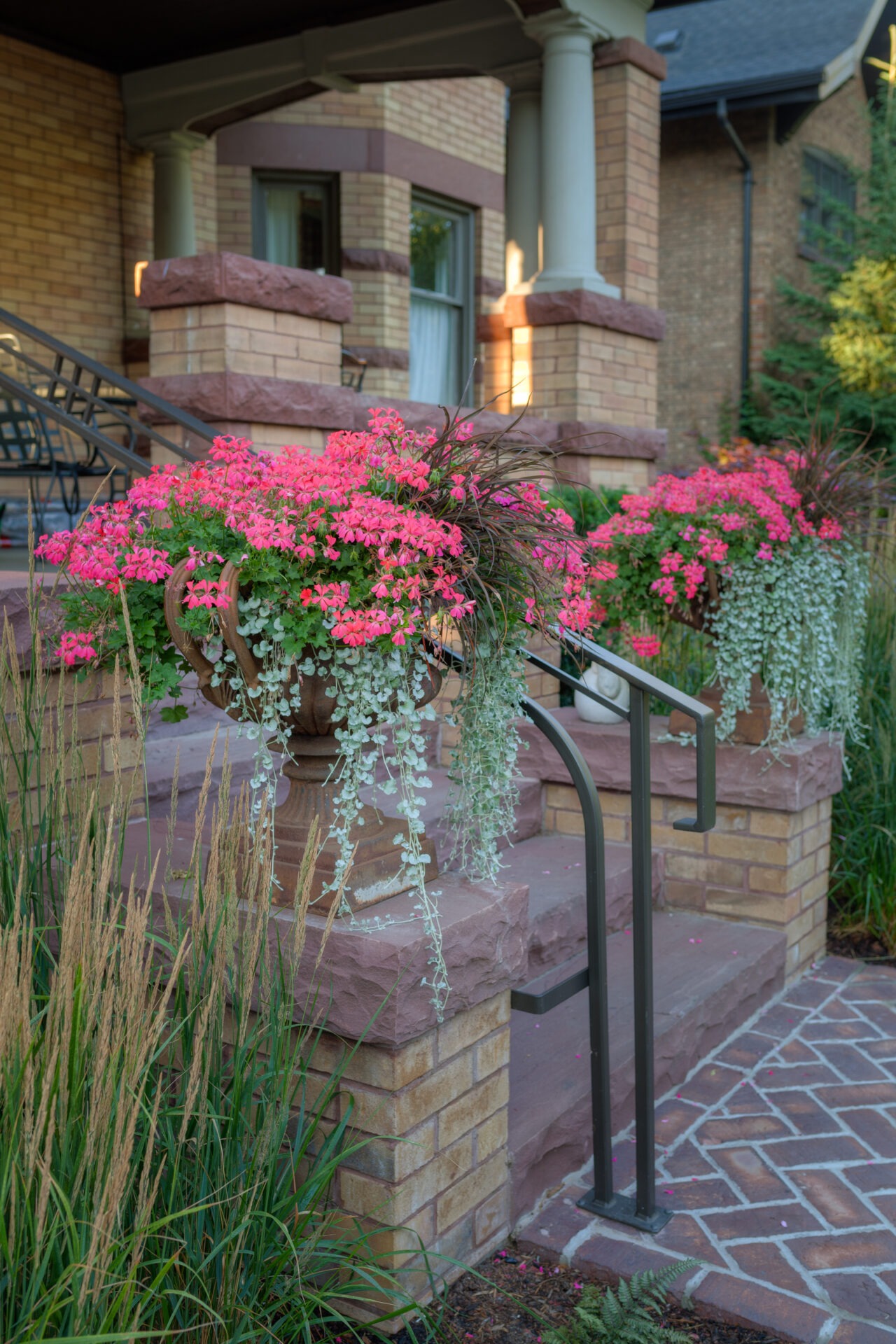 A cozy porch with ornate planters displays vibrant pink flowers, framed by brick columns, railings, and a patterned stone walkway.