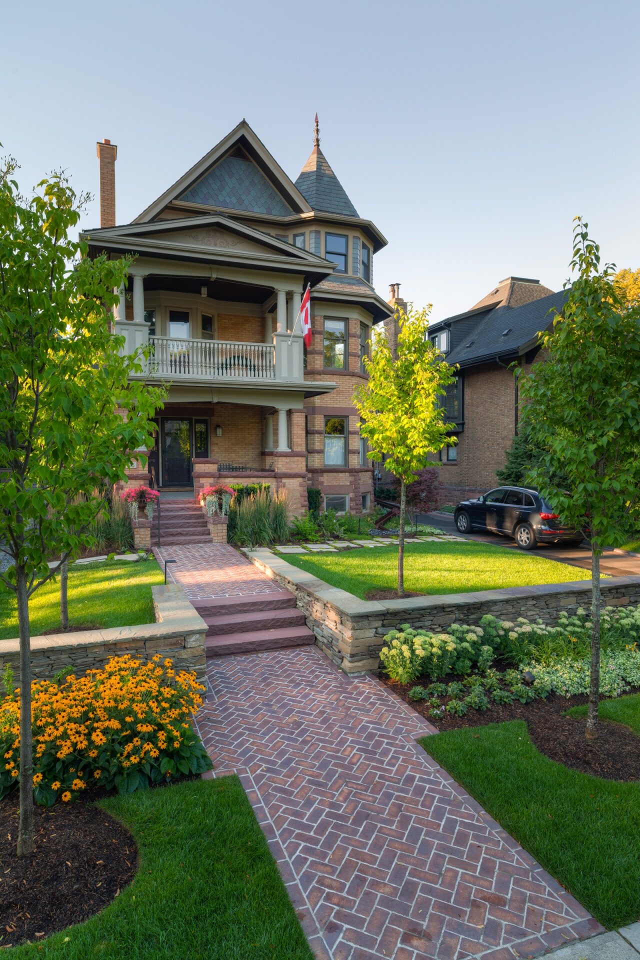 Victorian-style house with a Canadian flag, brick path, lush green garden, vibrant flowers, and parked car in a well-maintained residential neighborhood.