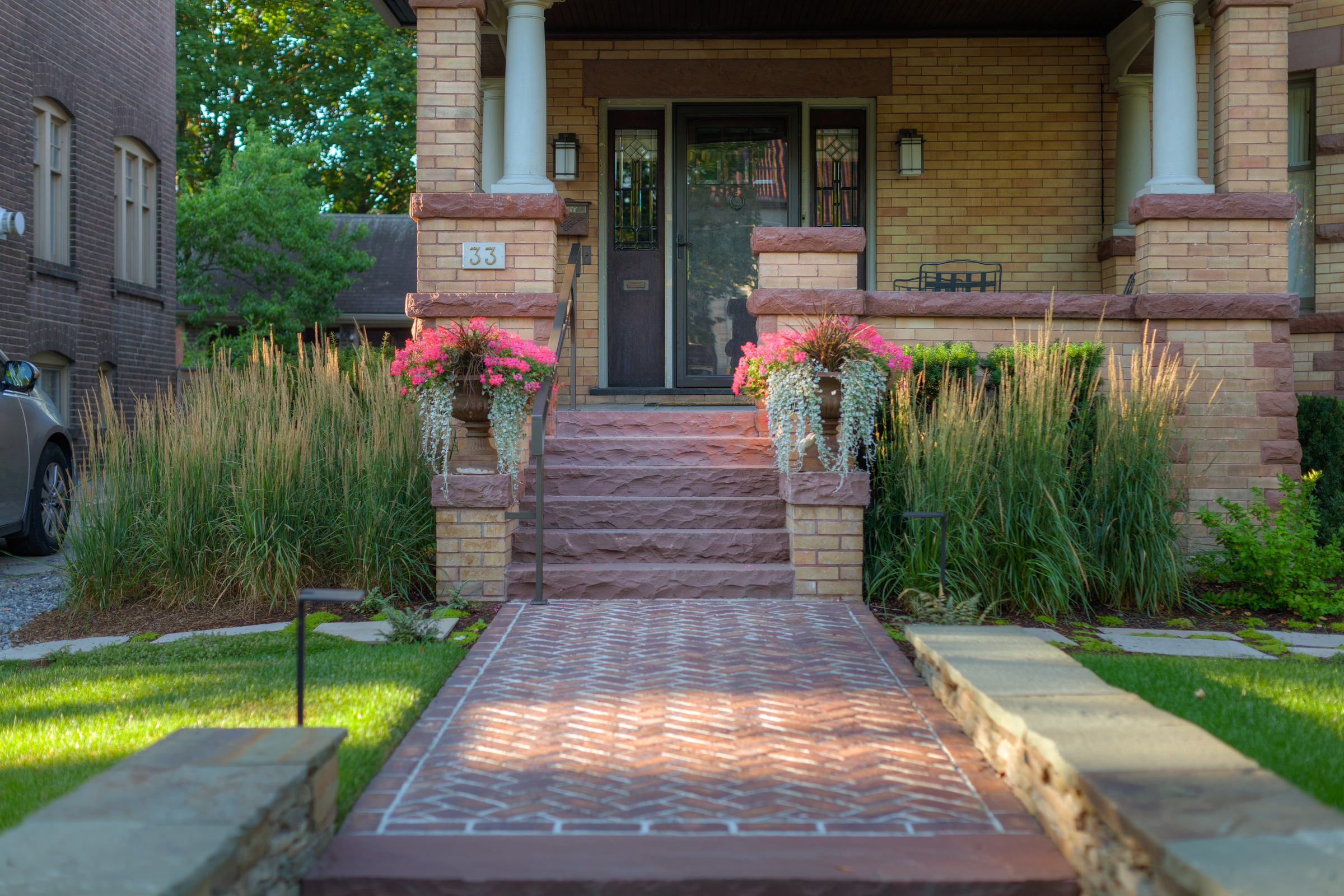 A charming brick house entrance with pink flower pots, stone steps, and a manicured lawn, surrounded by greenery and a parked car.