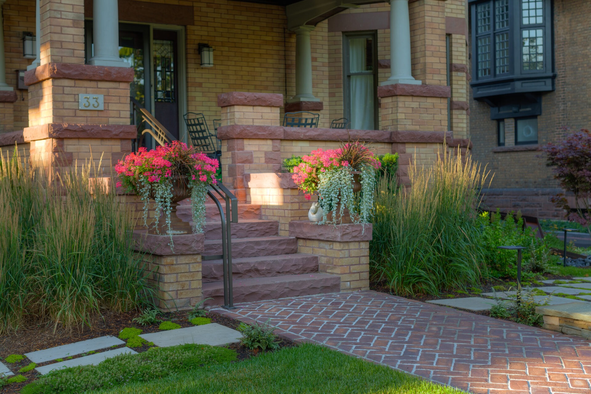 Charming brick house entrance with stone steps, vibrant flowers, and decorative planters. Well-maintained garden surrounds the inviting porch area.