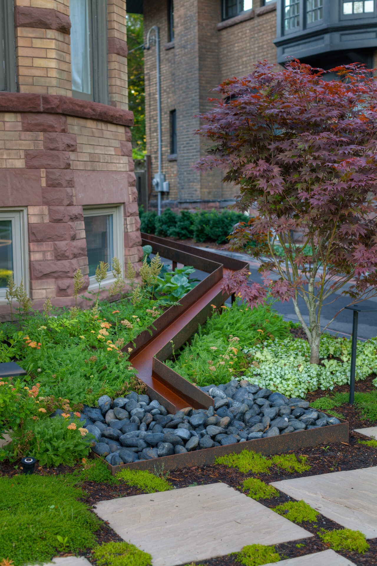 Brick building with landscaped garden, featuring a small tree, green plants, stepping stones, and a metal water feature over smooth rocks.