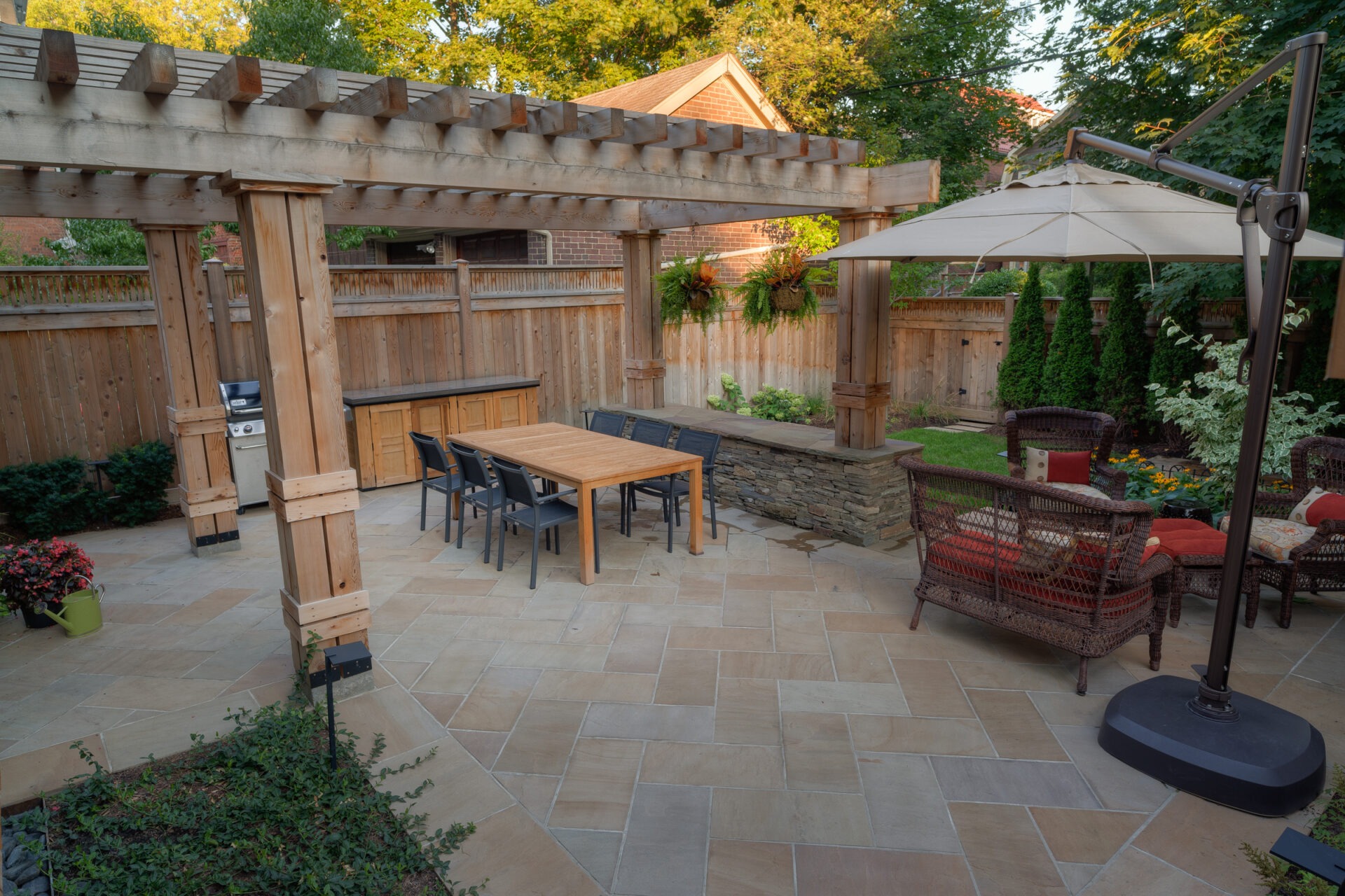 Outdoor patio with pergola, wooden dining table, black chairs, wicker furniture, and umbrella. Surrounded by a wooden fence and greenery.