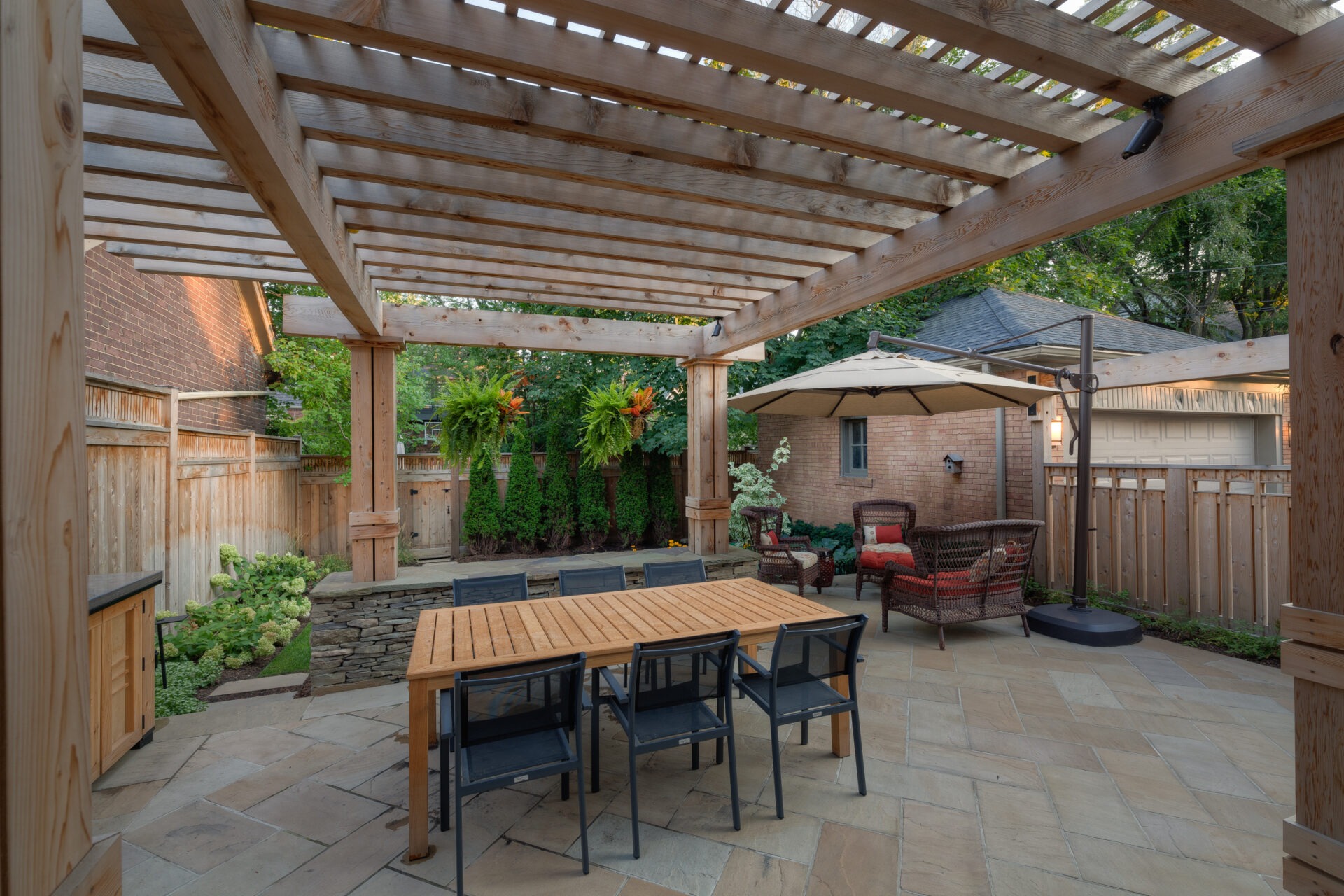 A backyard patio with a wooden pergola, outdoor dining set, comfortable seating, umbrella, and greenery in a private, fenced residential area.