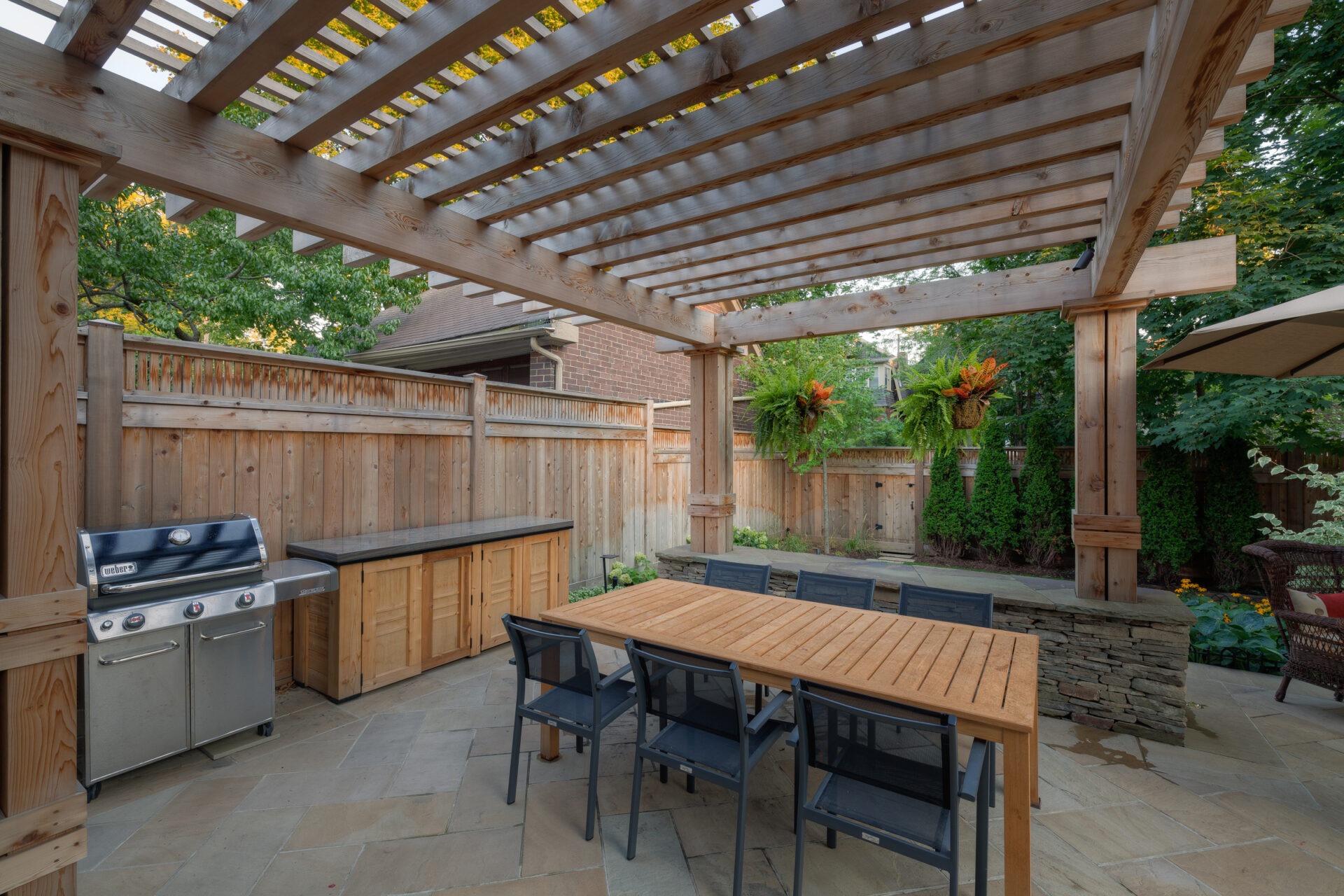 Wooden pergola over an outdoor patio area with dining table, chairs, and barbecue grill, surrounded by greenery and wooden fencing.