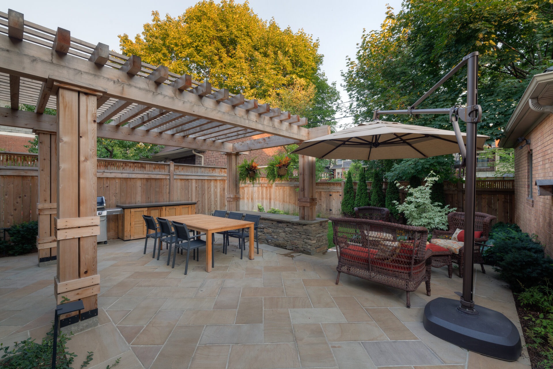 A cozy backyard patio features a pergola, outdoor dining set, wicker seating, umbrella, and lush greenery against a wooden fence backdrop.