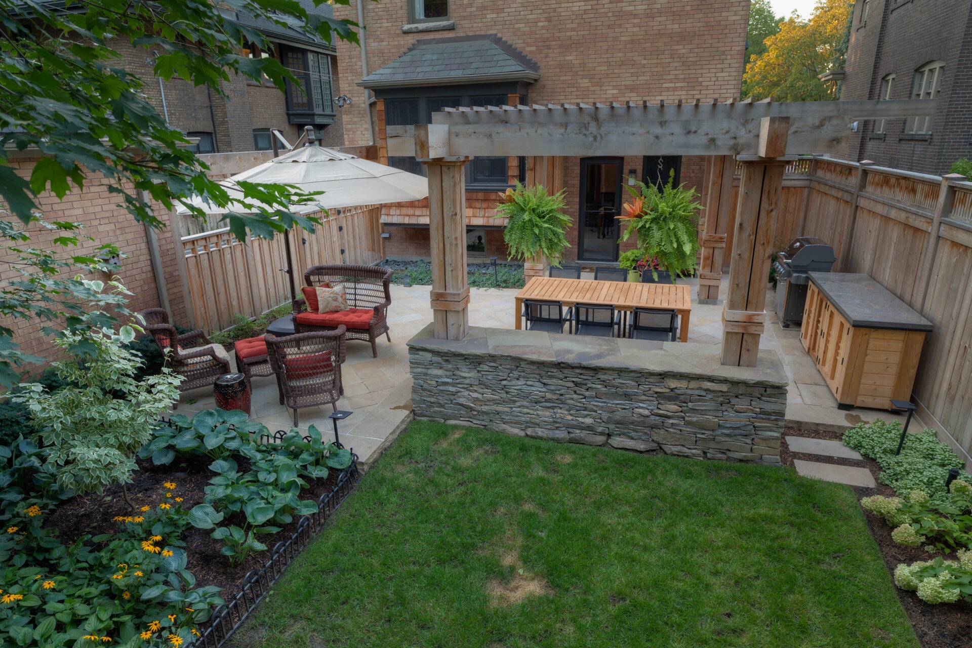 A cozy backyard patio features wicker furniture, an umbrella, wooden pergola, dining table, grill, vibrant plants, and a small garden area.