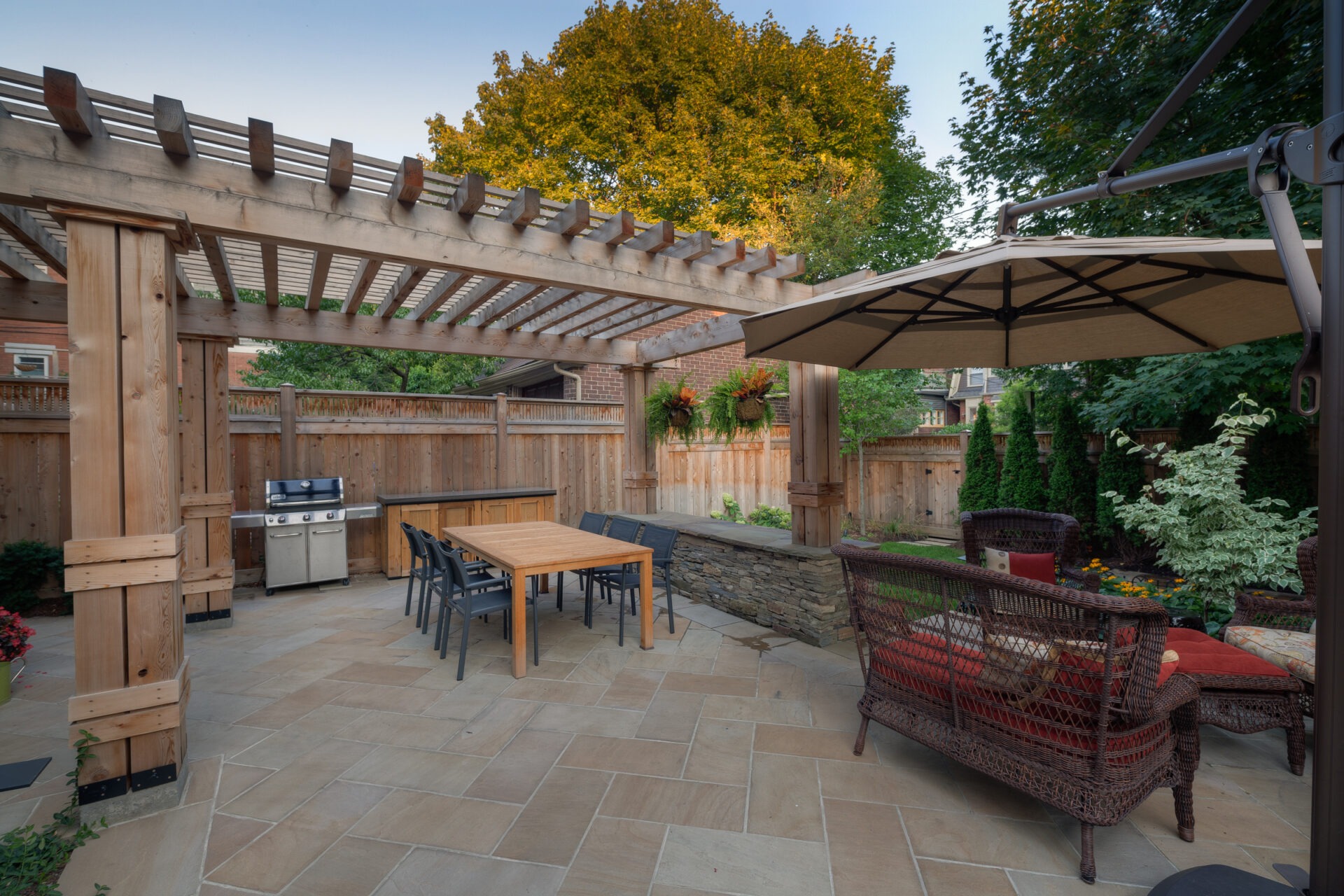 Outdoor patio with pergola, table, chairs, wicker furniture, and grill, surrounded by plants and trees, creating a cozy garden setting.