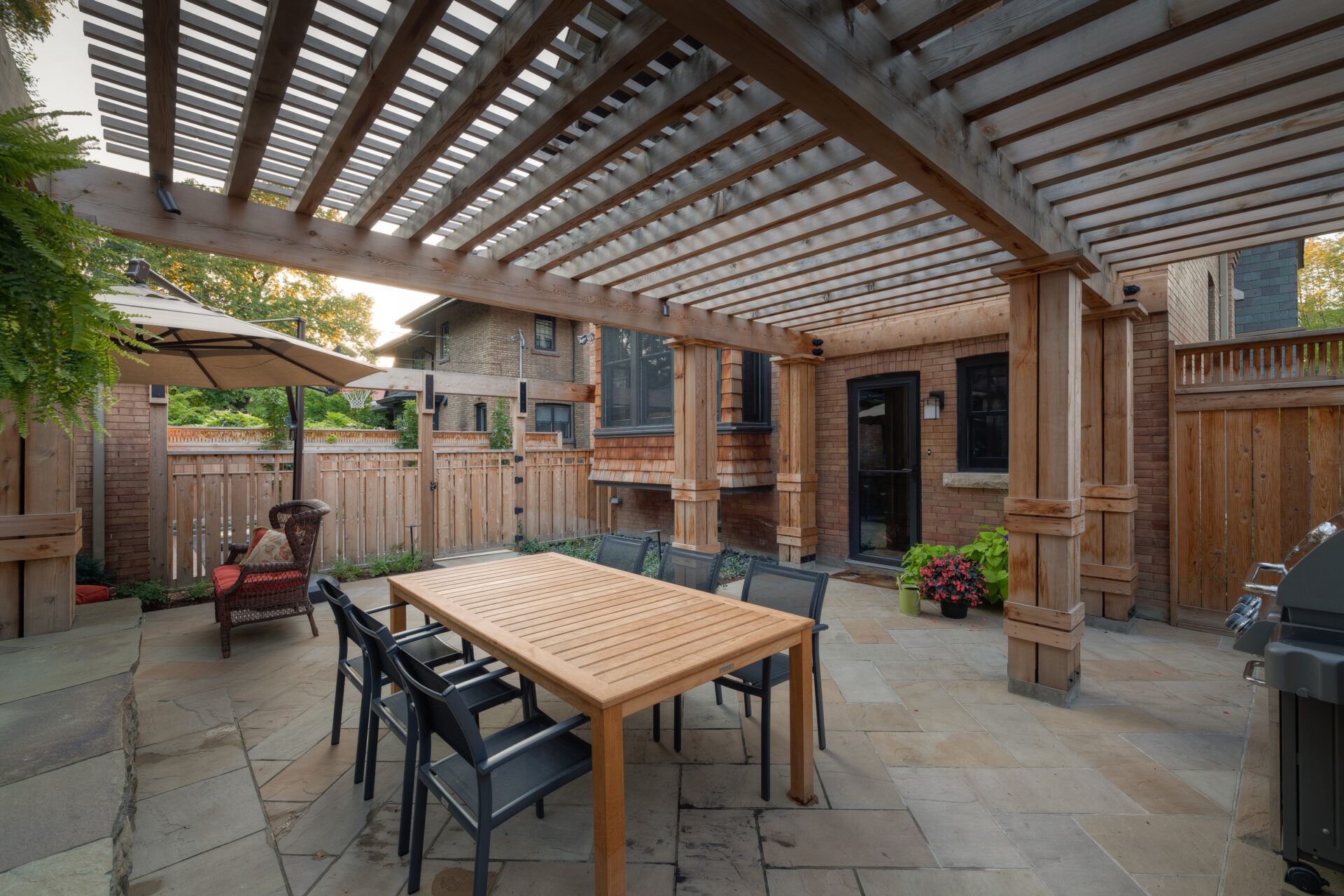 A wooden pergola shades a backyard patio with a table, chairs, umbrella, and grill. Fenced garden area with plants and flowers.