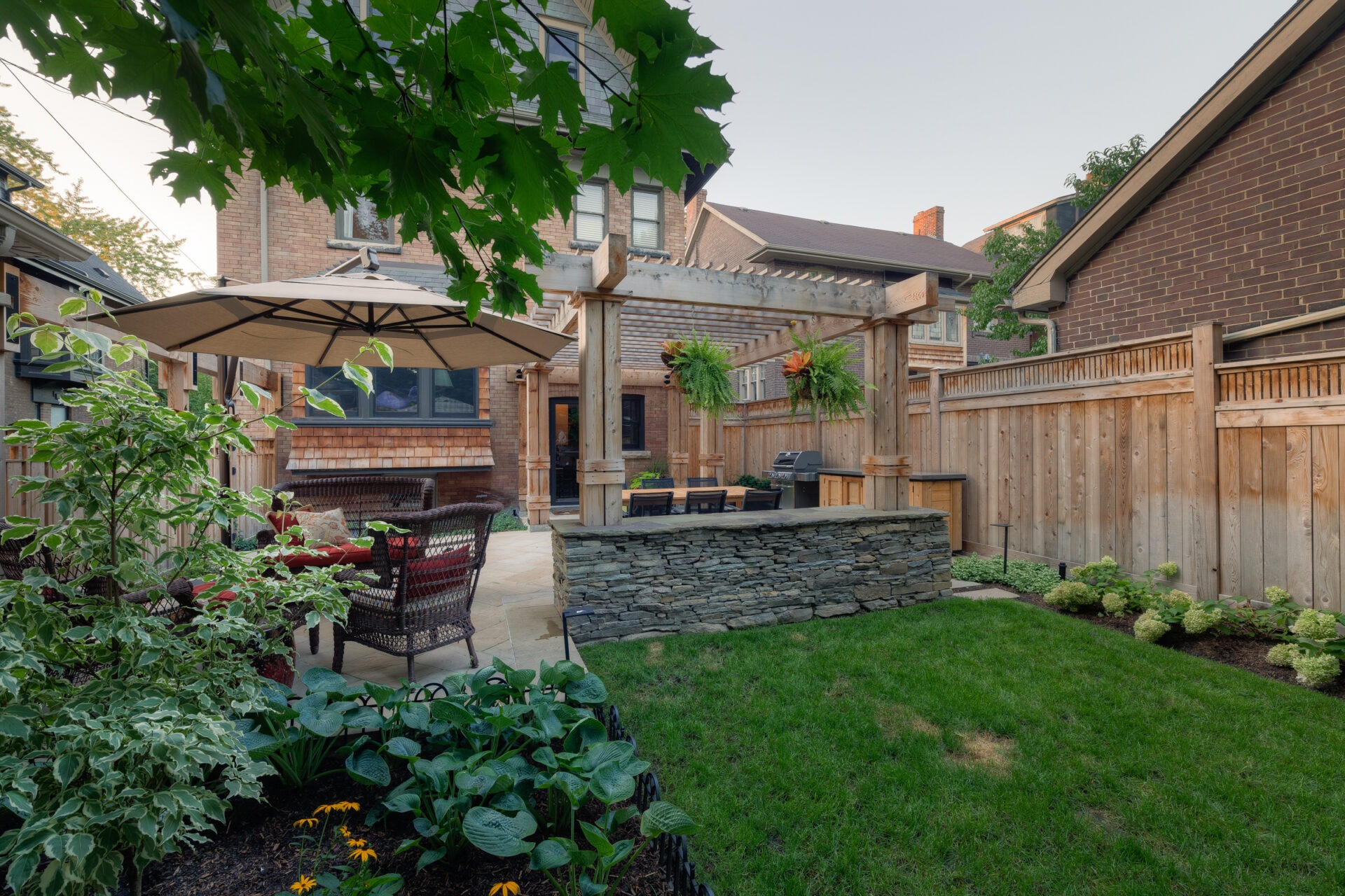 A backyard garden features a pergola, patio furniture, and a barbecue grill, surrounded by greenery and mature trees beside a brick house.