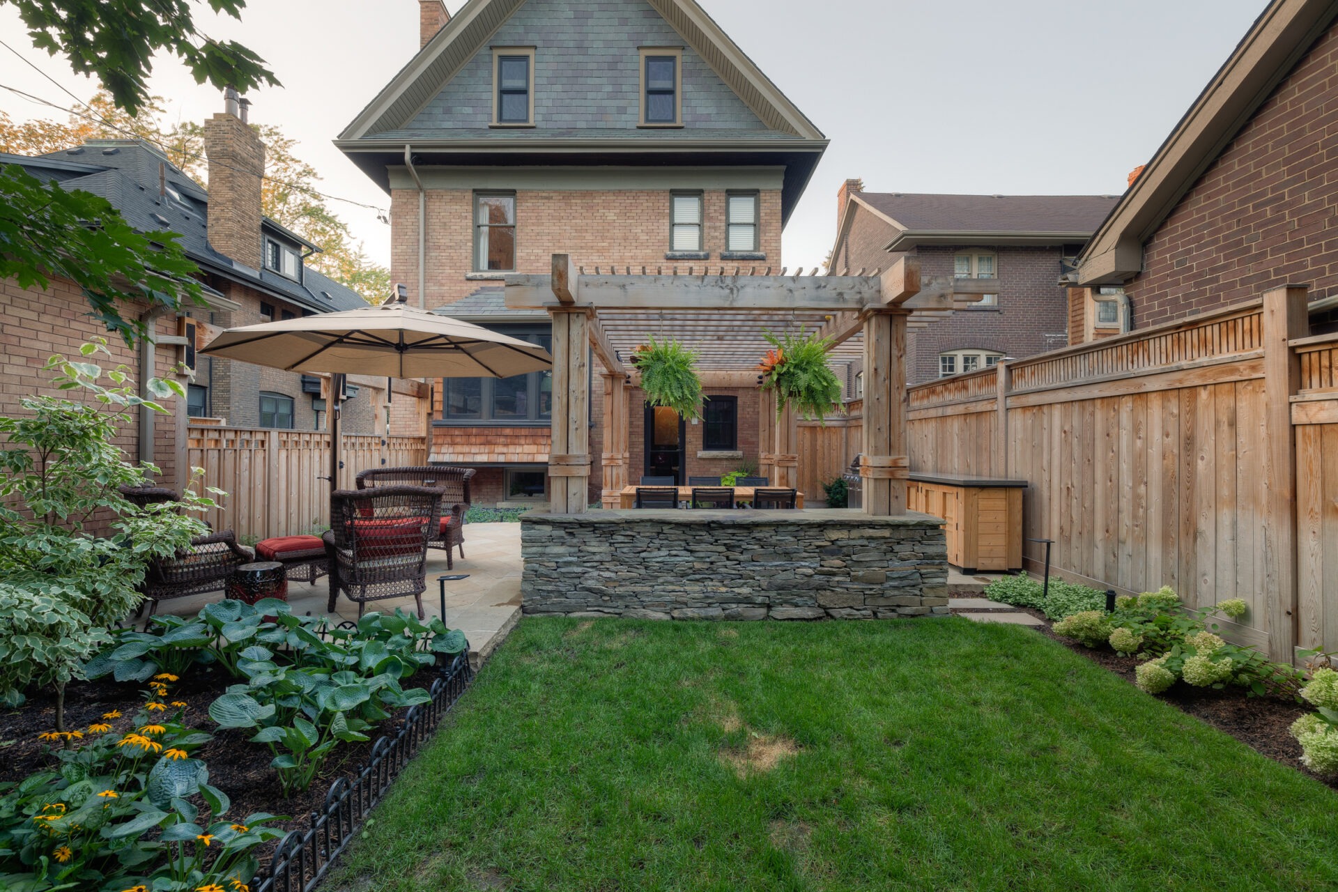 A charming backyard features a wooden pergola, patio furniture, lush garden, and a traditional brick house surrounded by a tall fence on a sunny day.