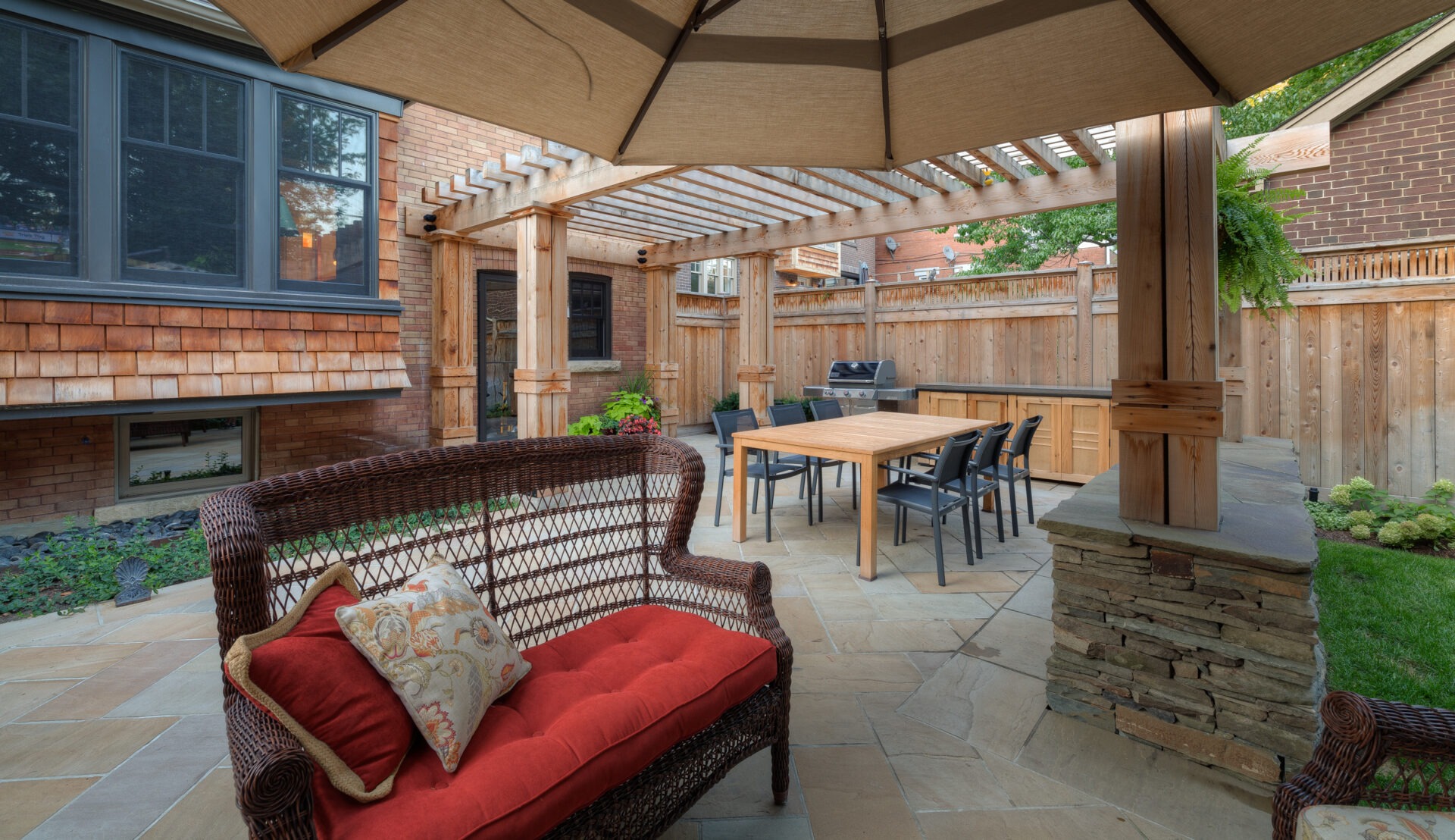 Cozy backyard patio with wicker furniture, red cushions, wooden pergola, barbecue grill, dining table, and lush greenery surrounded by a wooden fence.