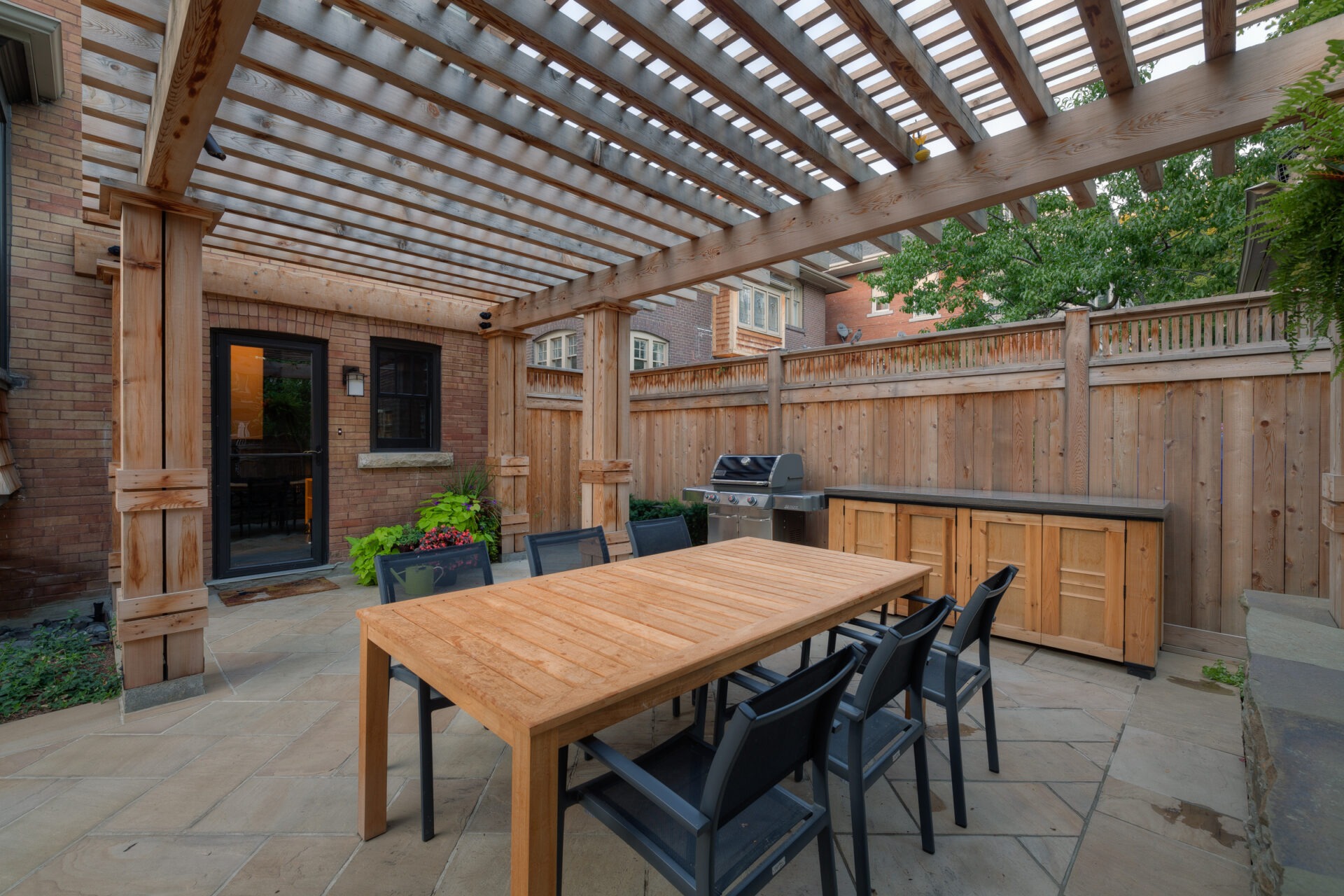 Wooden pergola covers patio with dining table, chairs, and barbecue grill. Enclosed by wooden fence, brick exterior, and plants visible.