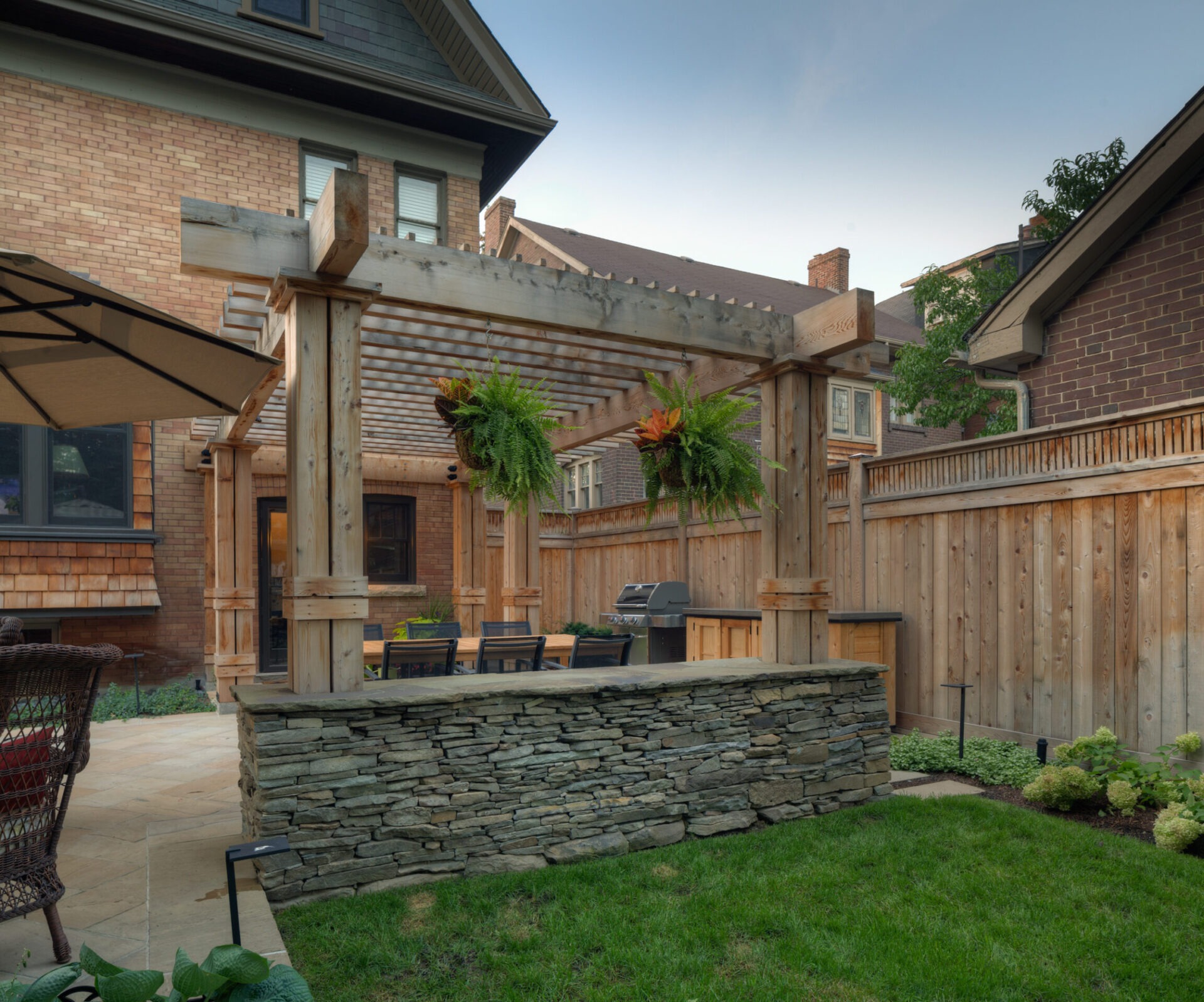 A charming backyard features a wooden pergola with hanging plants, stone wall, patio furniture, and a barbecue, surrounded by a wooden fence.