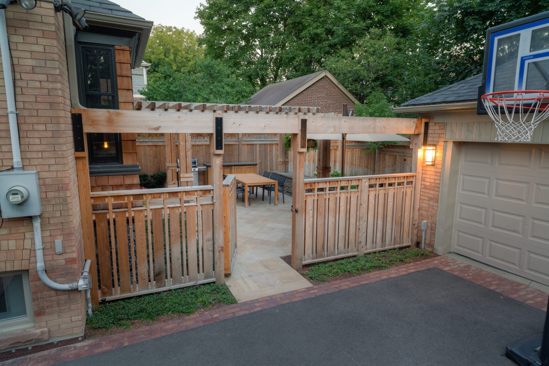 A cozy backyard patio with wooden pergola, table, and chairs, set against a brick house. Basketball hoop on garage, surrounded by trees.