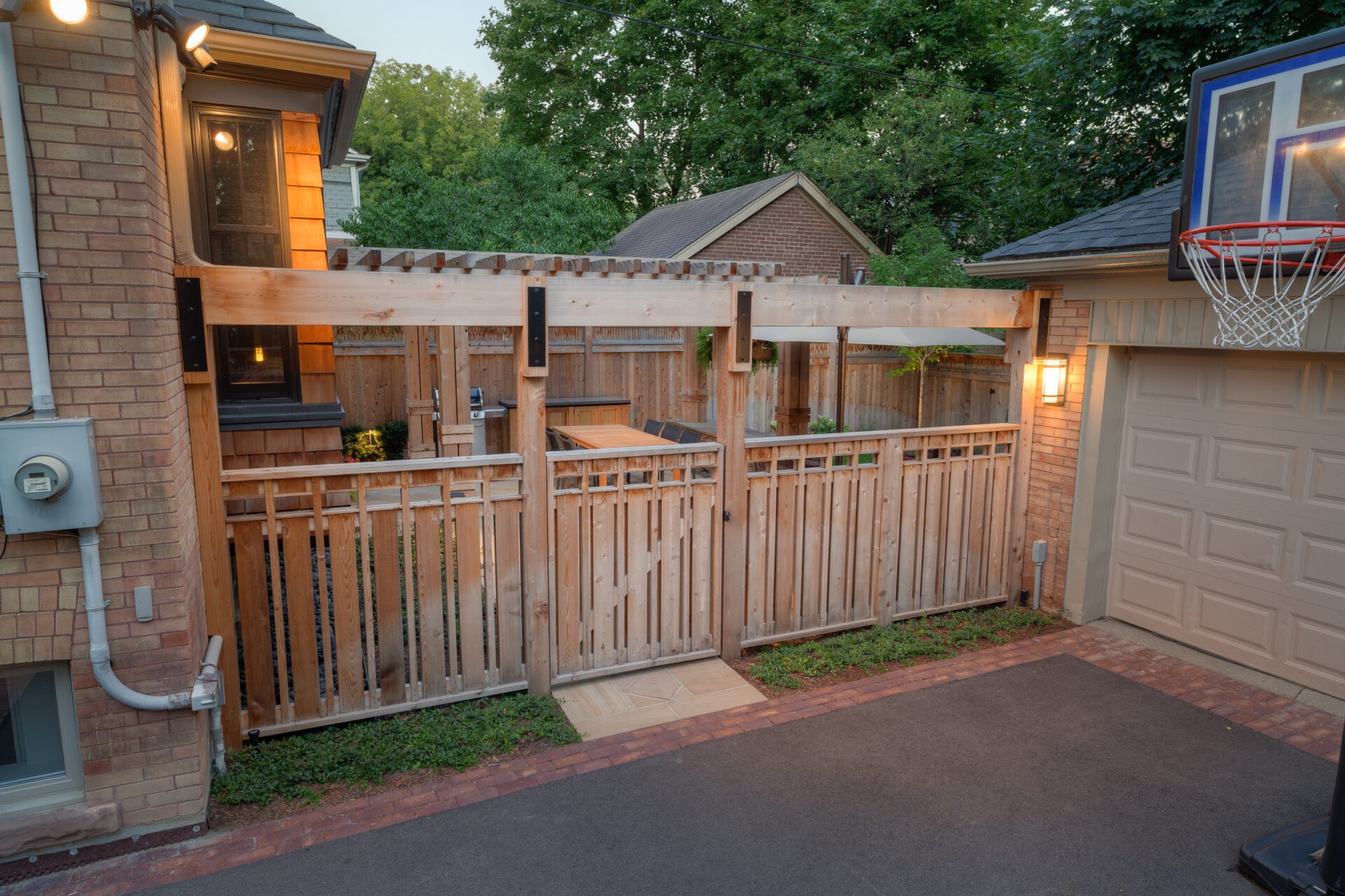 A backyard scene featuring wooden fencing, a pergola, and a basketball hoop. There's a garage, green foliage, and exterior lighting.