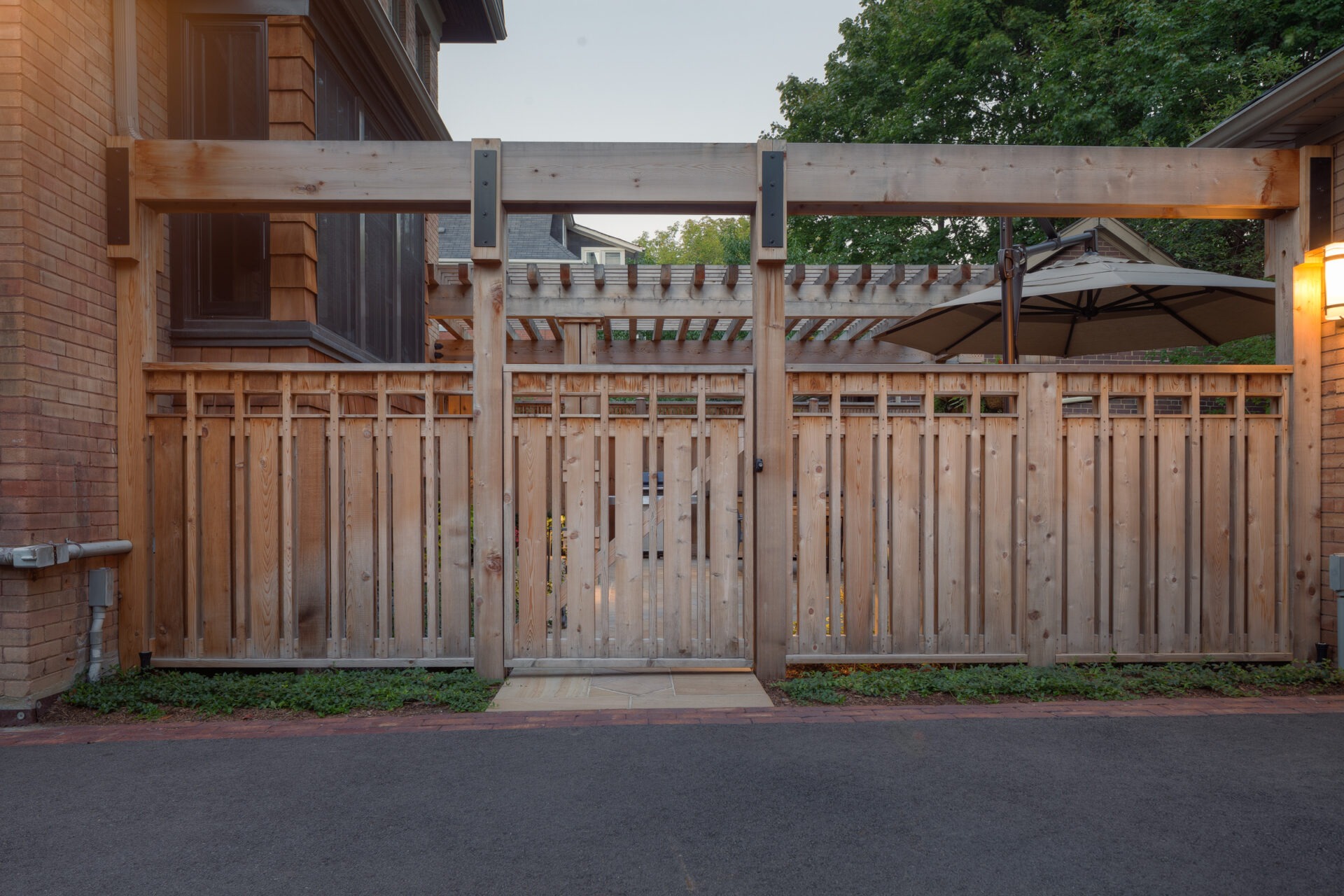 A wooden gate and fence next to a brick building, surrounded by greenery. A large umbrella is partially visible behind.