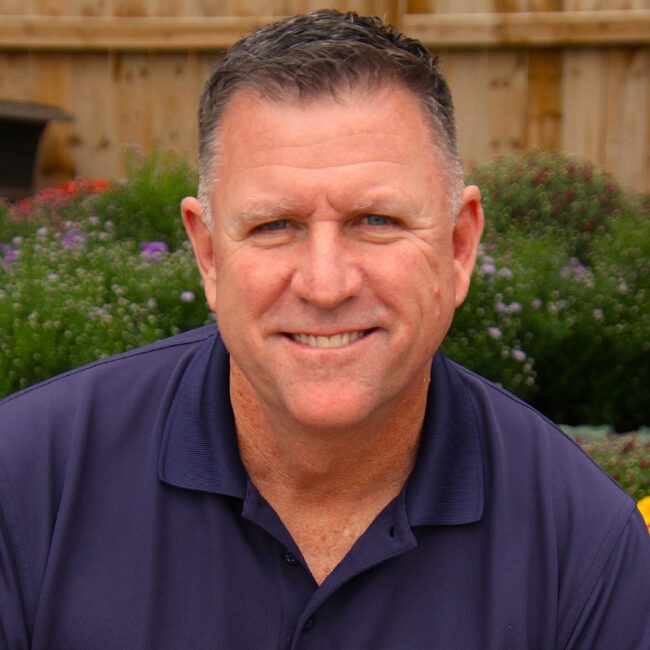 A person in a navy blue shirt smiles at the camera, with green foliage and flowers in the background.