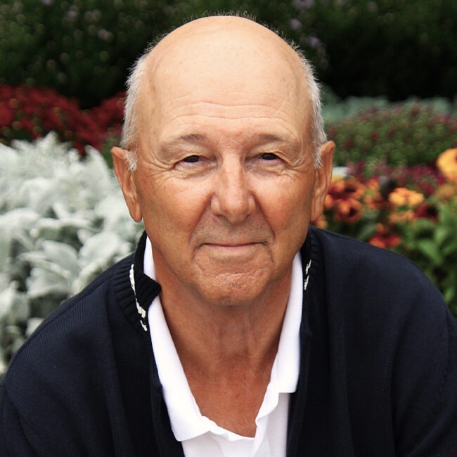 A person with a warm expression stands before lush foliage and vibrant flowers, creating a serene and natural backdrop.